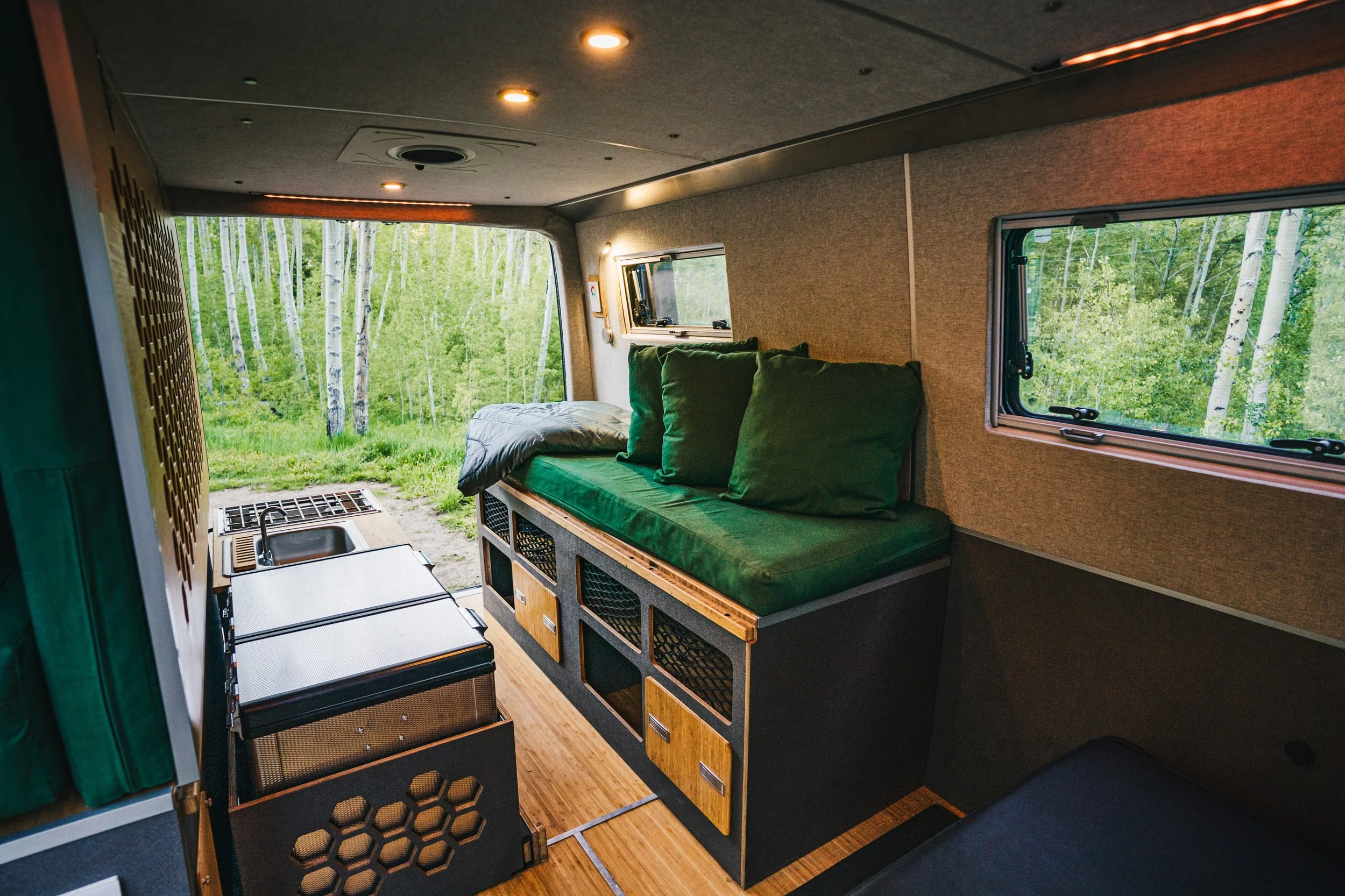 Interior of a camper van with a built-in bed with green bedding and pillows, window with a forest view, small kitchen area with a sink and countertop, and wood accents.