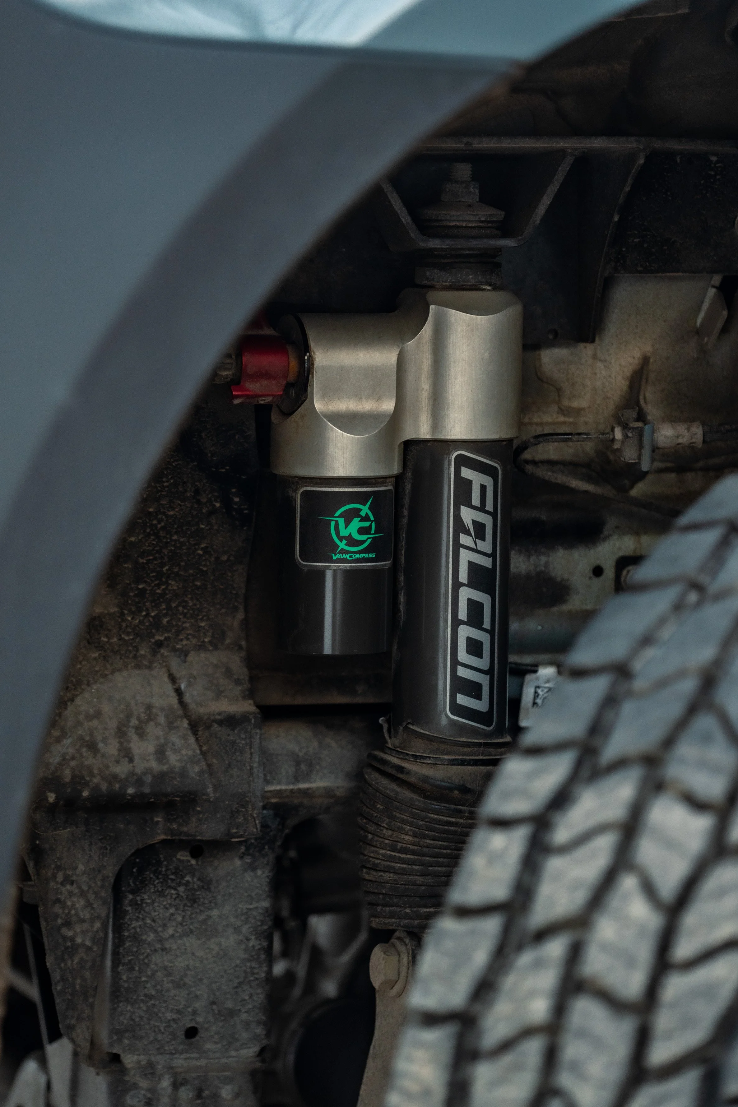 Close-up of a vehicle's suspension system, featuring a Falcon shock absorber and a steering stabilizer, with part of the tire and wheel well visible.