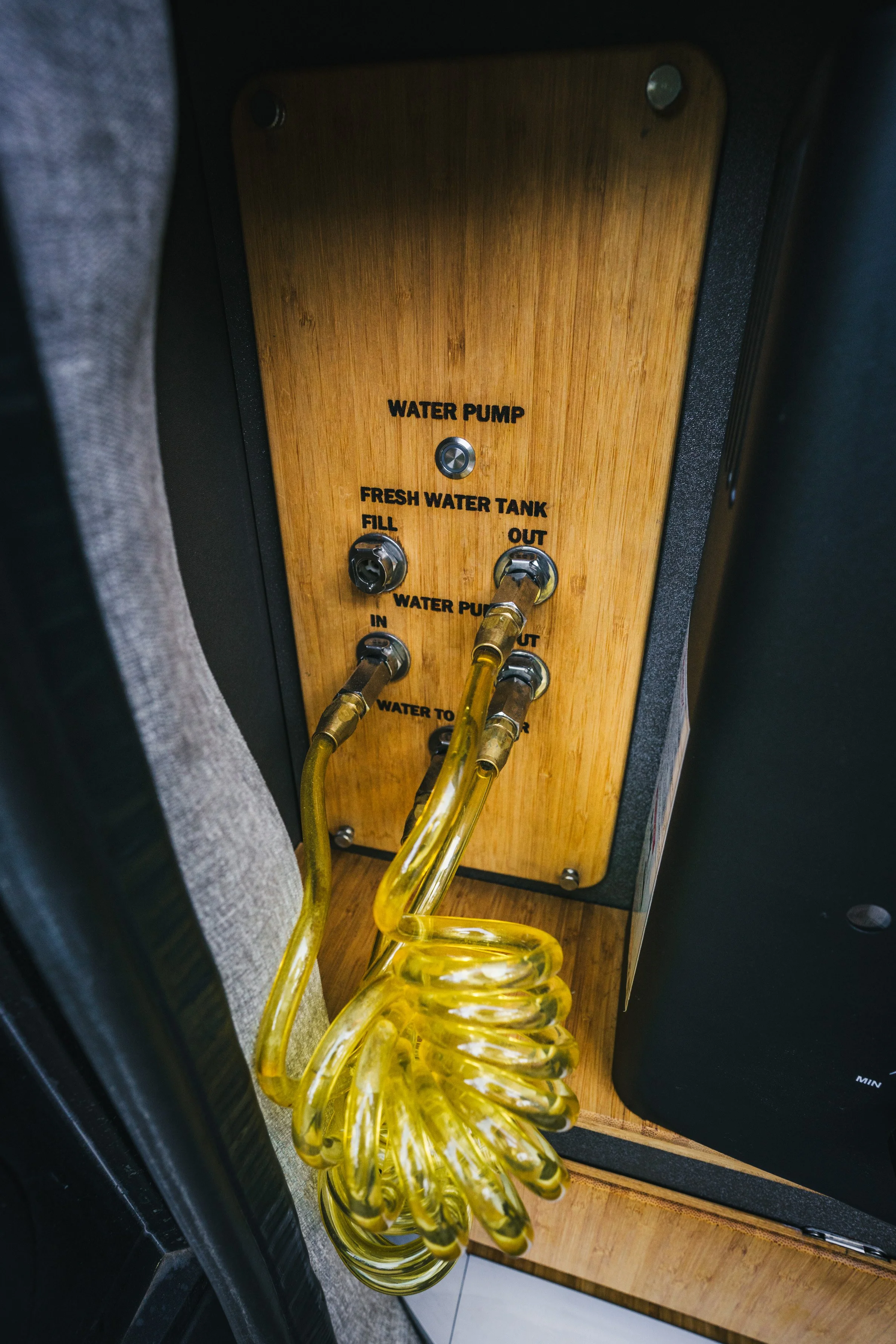 A wooden panel with water pump controls, including labels for Fresh Water Tank Fill, Water Pump In/Out, and Water to Heater, with yellow flexible tubing connected to the water pump.