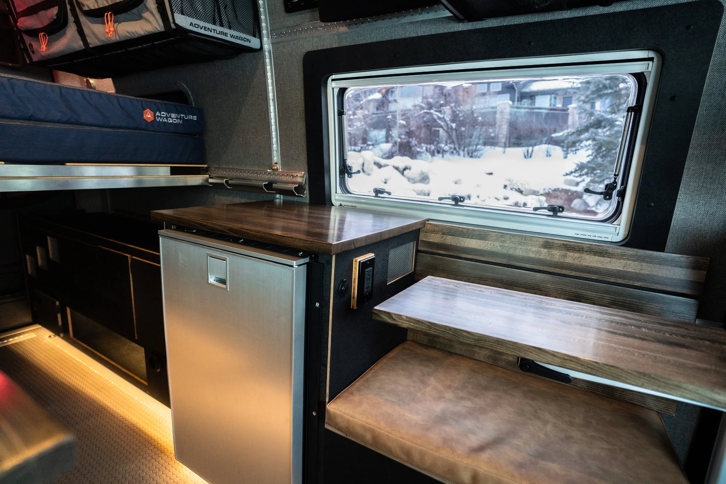 Interior of a camper van with a small refrigerator, a wooden table, bench seating with a brown cushion, and a window showing snowy landscape outside.