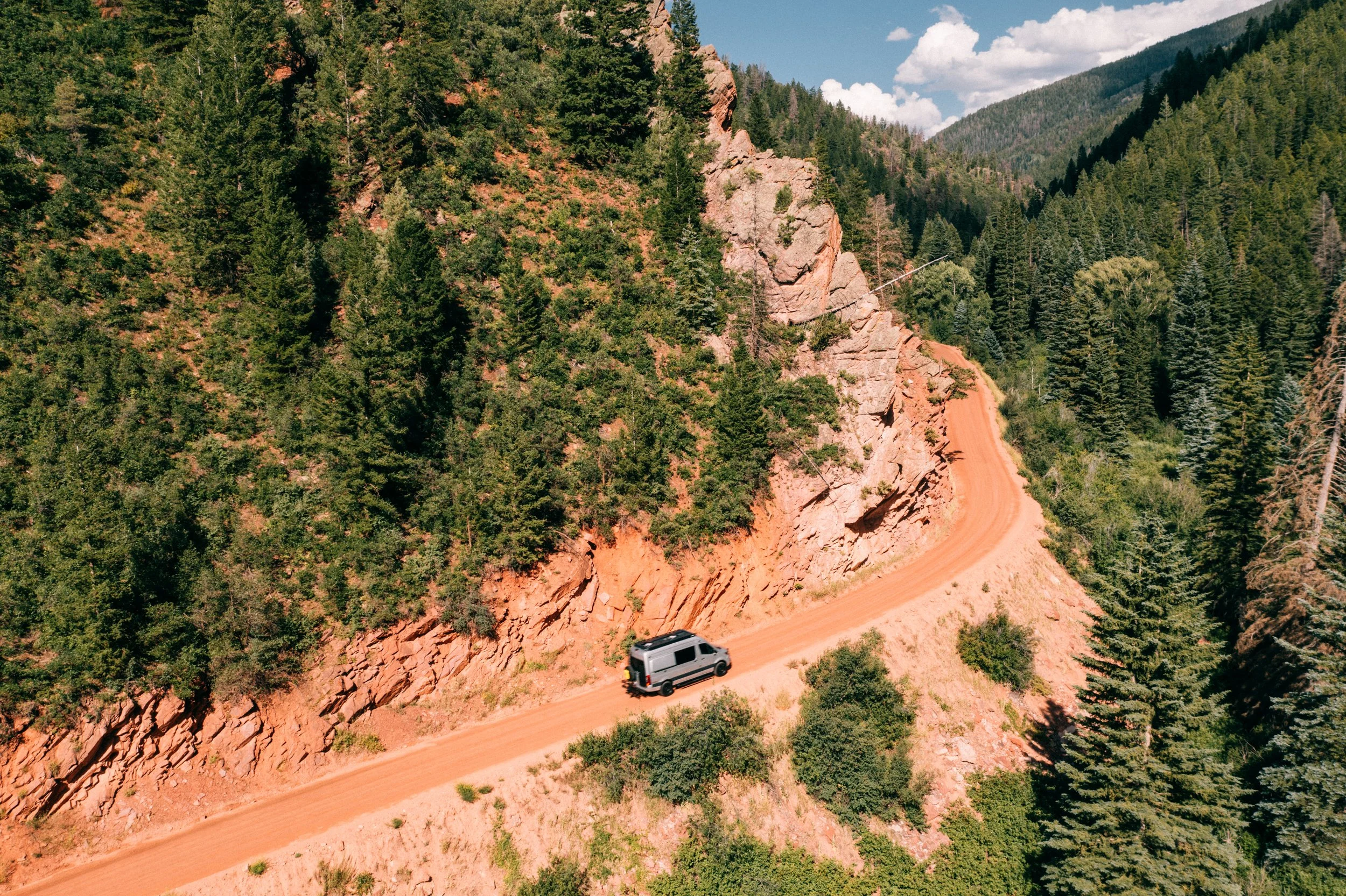 A black van driving on a winding dirt road surrounded by a forest of tall green trees and rocky hills under a partly cloudy sky.