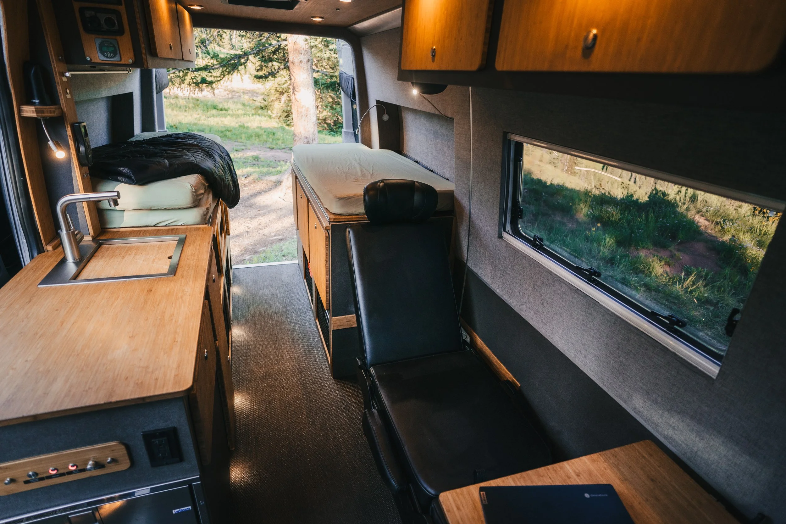 Interior of a camper van with wooden cabinetry, a small sink, a bed with bedding, a black chair, and windows showing greenery outside.