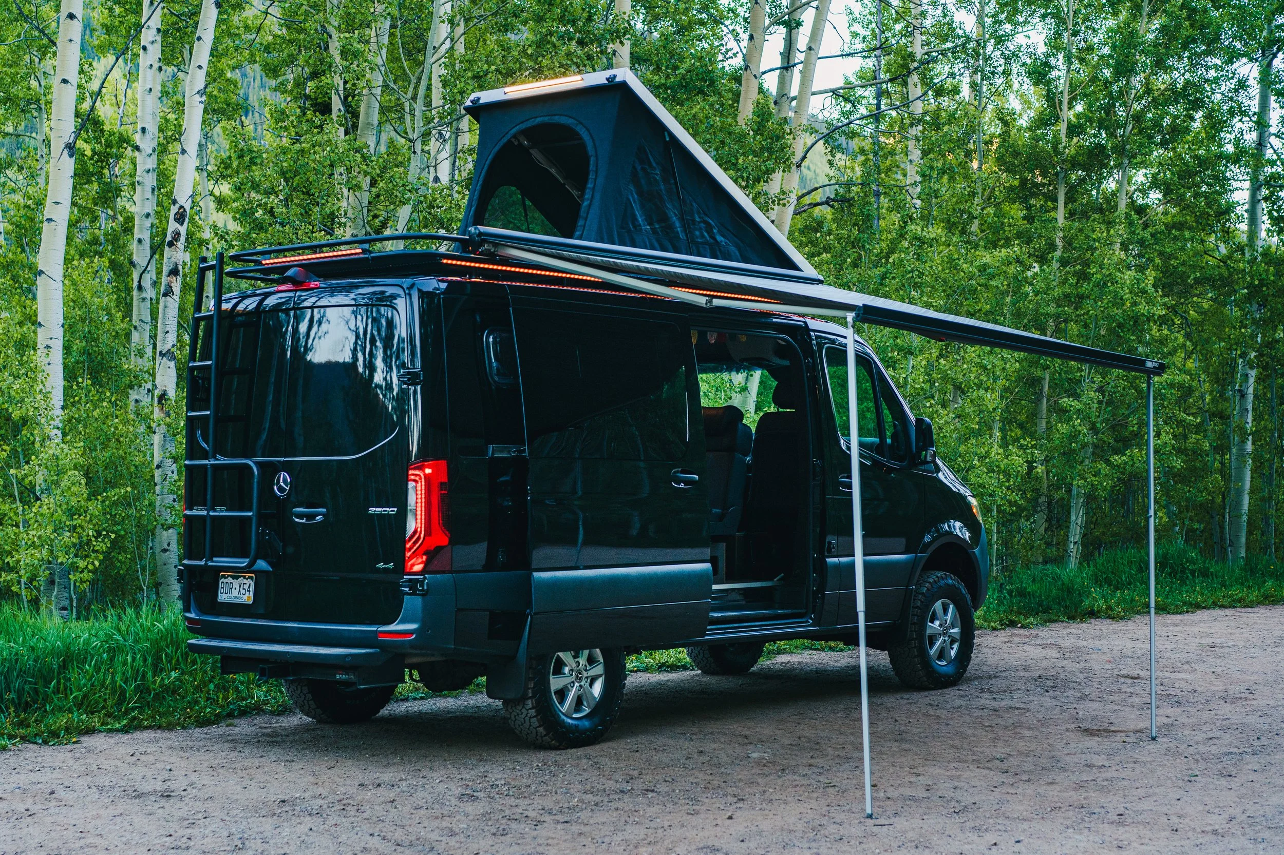Black camper van with extended roof tent and awning parked on dirt path in a forested area with green trees.