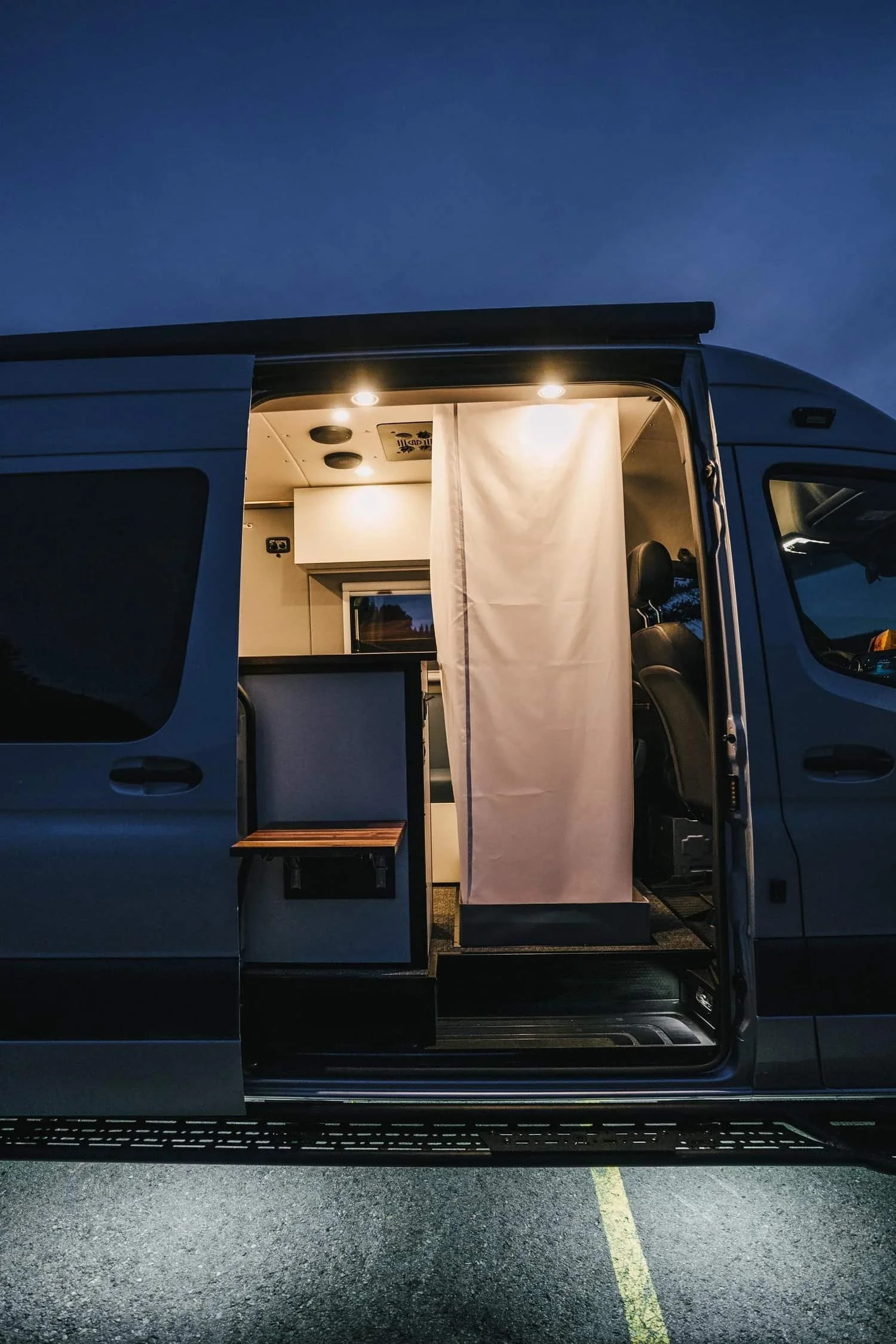 Interior of a converted camper van at dusk with lighting, a hidden shower, seating, and kitchen area.