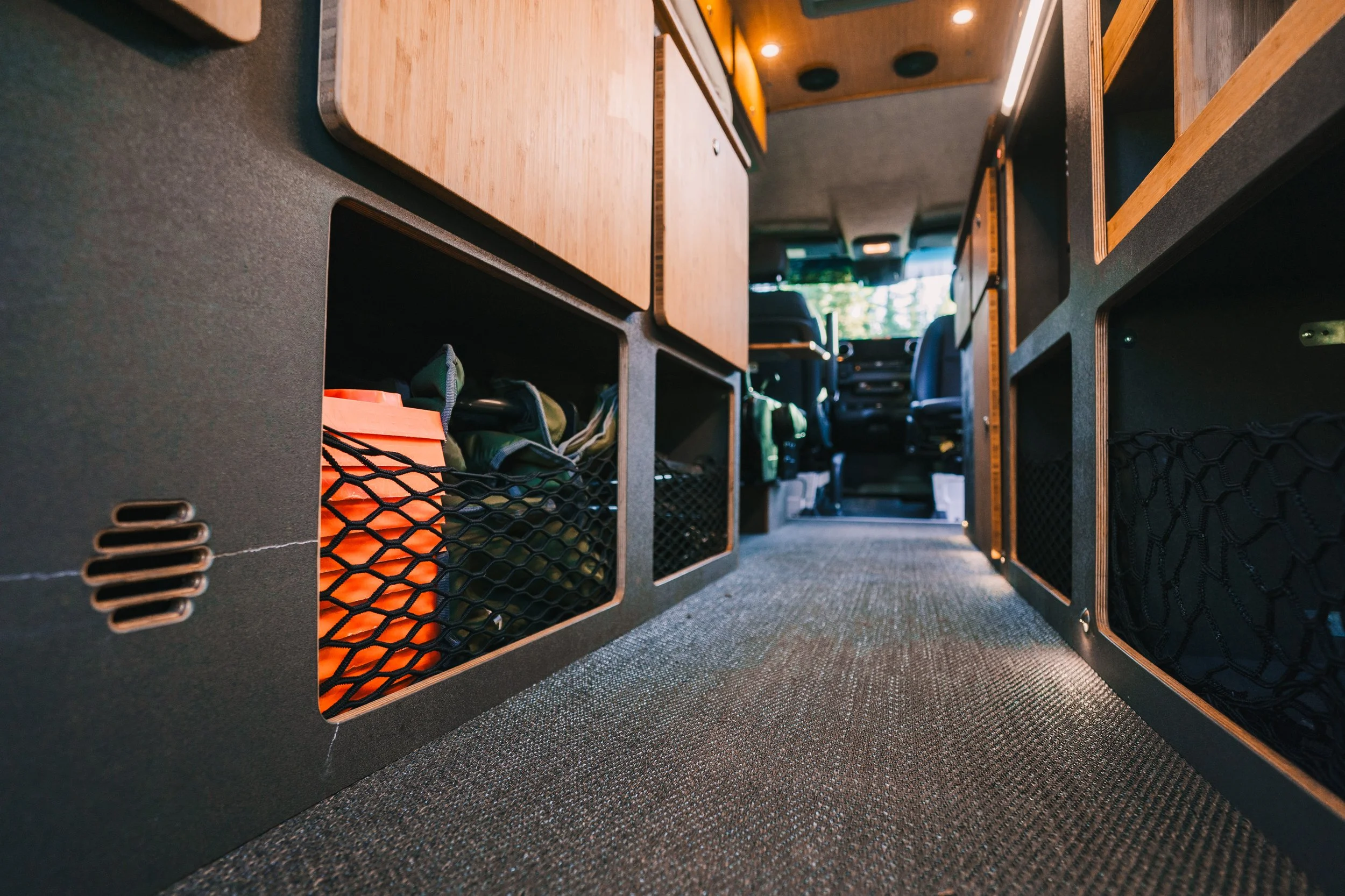 Inside a van or camper with storage compartments along the sides, showing various bags and items stored in black mesh pockets, with the dashboard and driver seats visible in the background.