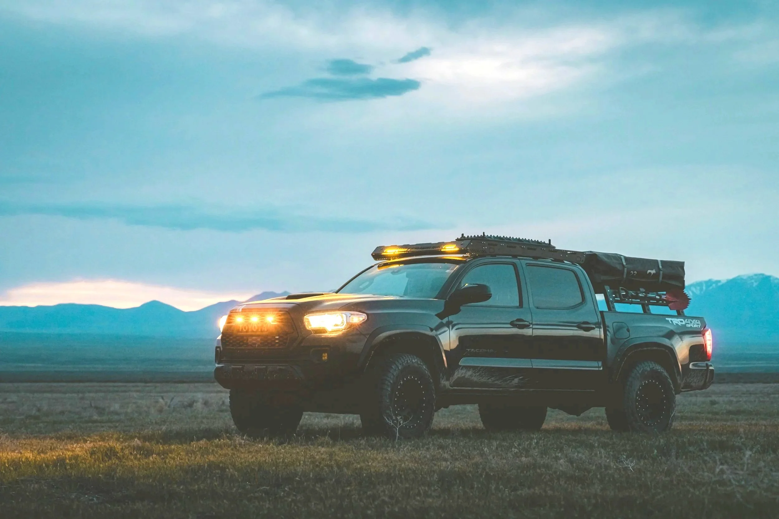 A black Toyota Tacoma pickup truck parked on a grassy field with mountains and a cloudy sky in the background during dusk or dawn.