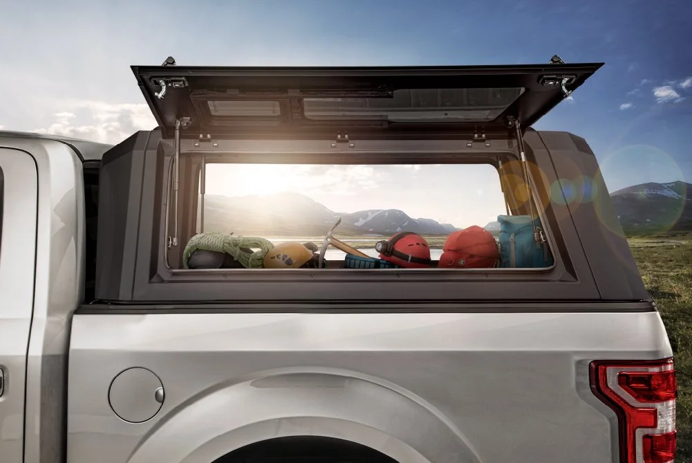 Back of a pickup truck with camping gear including helmets, a tent, and a bag, set against a mountain landscape with a partly cloudy sky.