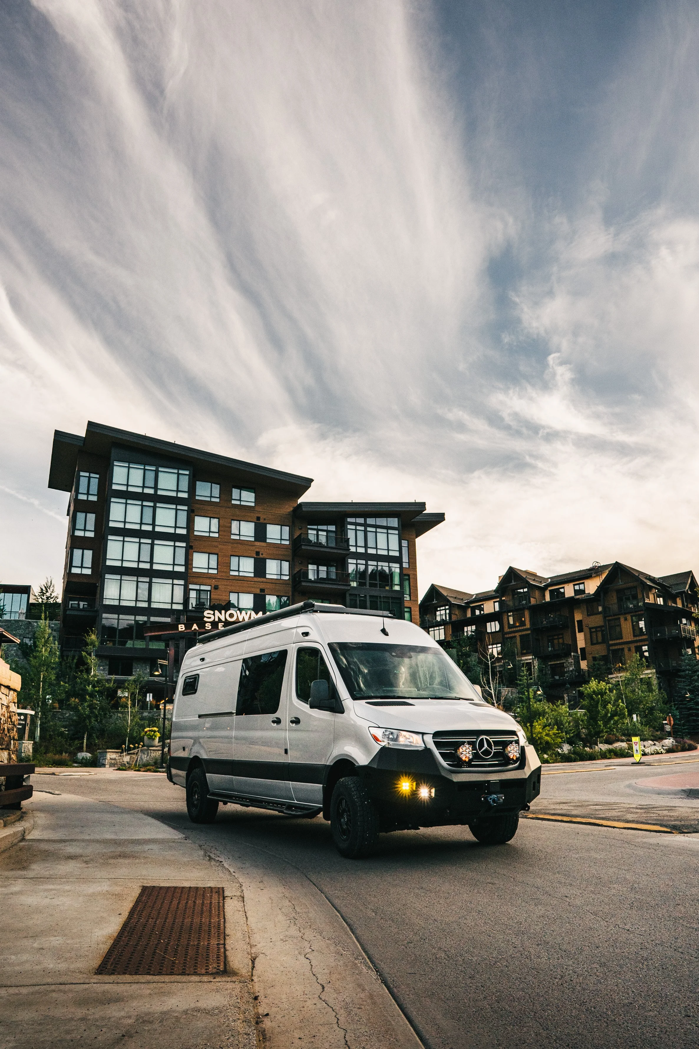 A silver Mercedes-Benz Sprinter van parked on a city street with modern apartment buildings and cloudy sky in the background.