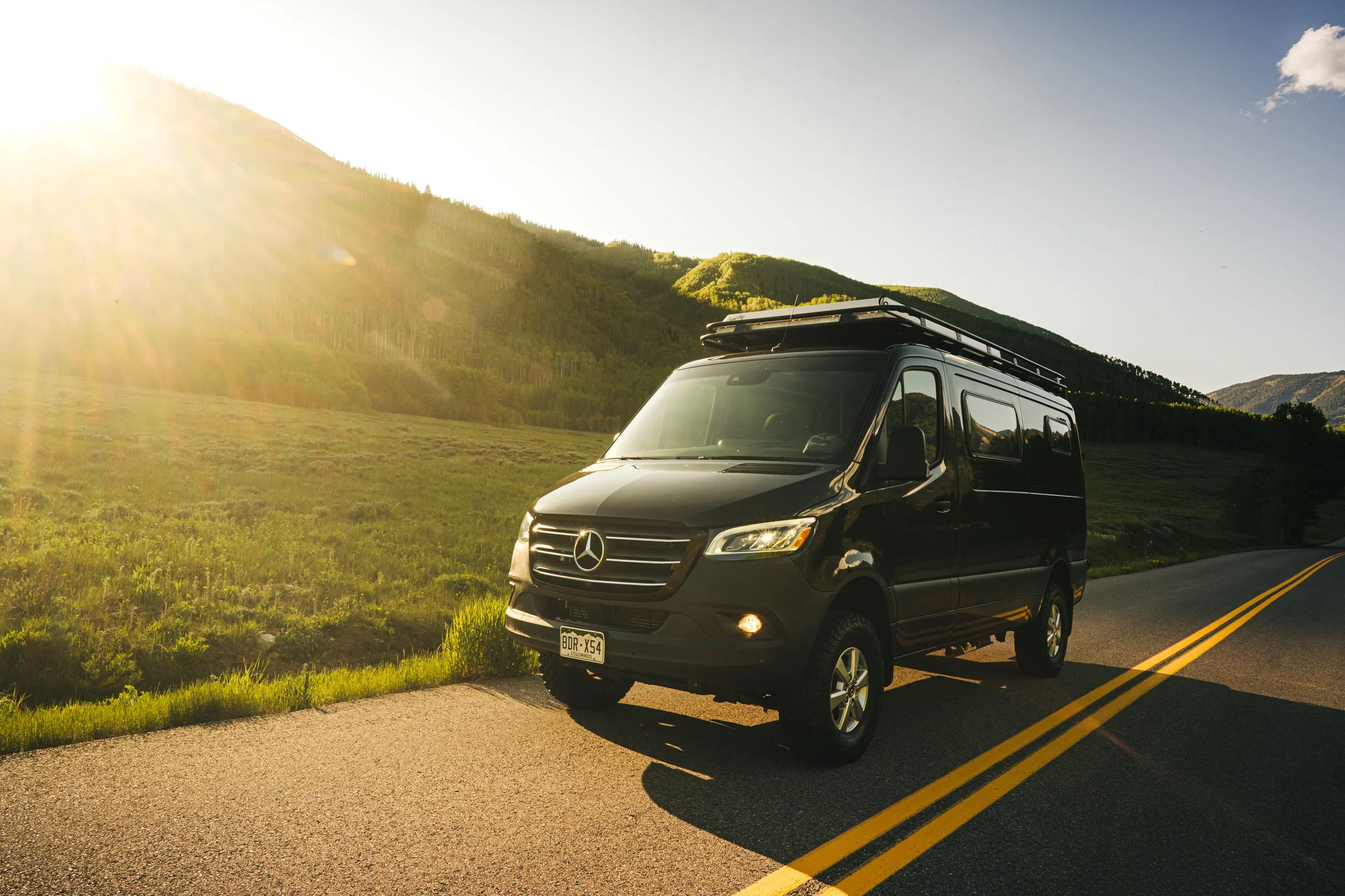 Black Mercedes-Benz camper van parked on the side of a rural road with green hills and mountains in the background, sunlight shining from the left.