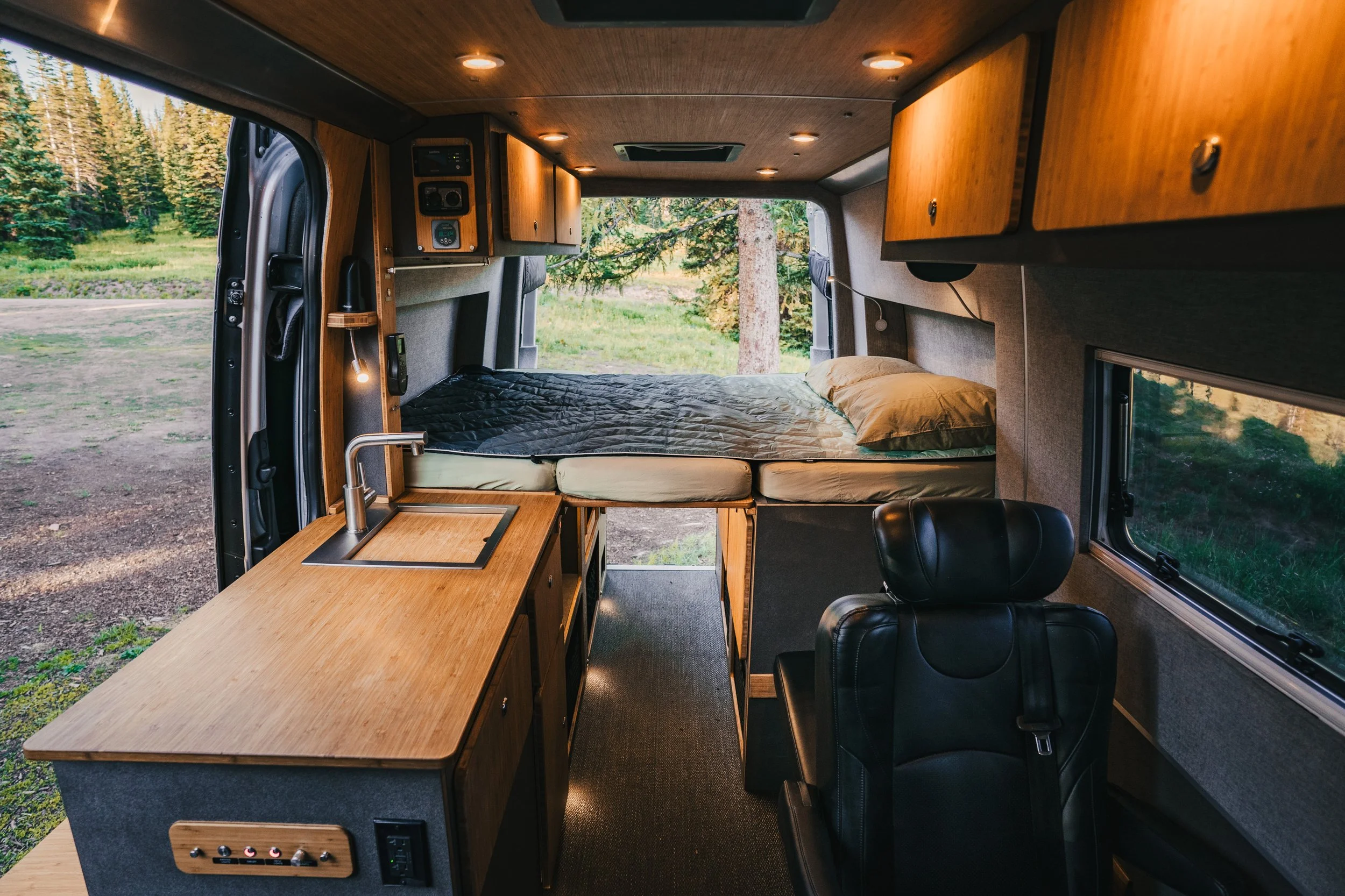 Interior of a converted van with a bed, small kitchen area, and window showing trees outside.