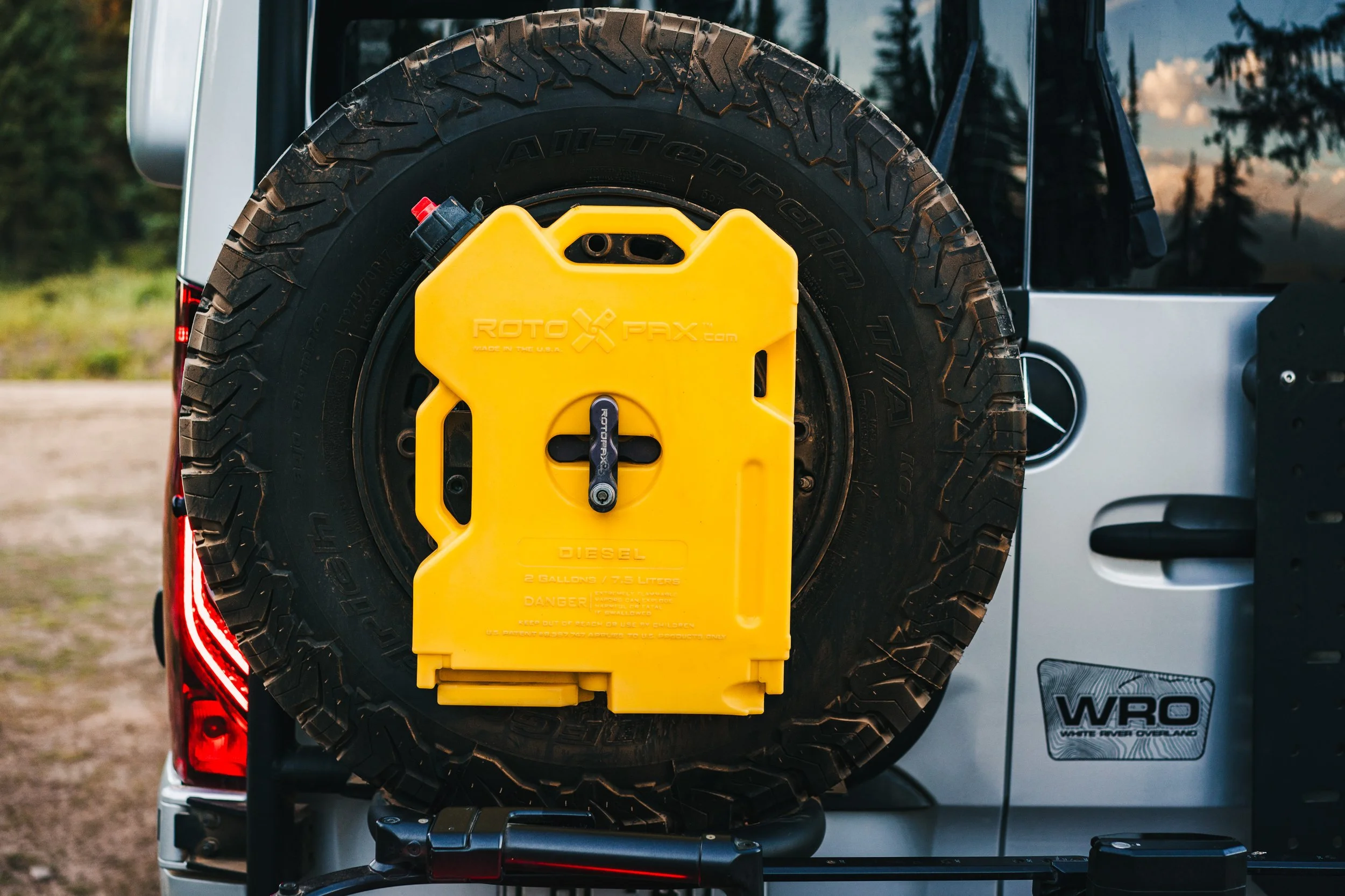Rear view of a silver vehicle with a spare tire mounted on the back. The tire has a yellow fuel transfer tank attached to it. The vehicle has a black door handle and a sticker that reads 'WRO'.