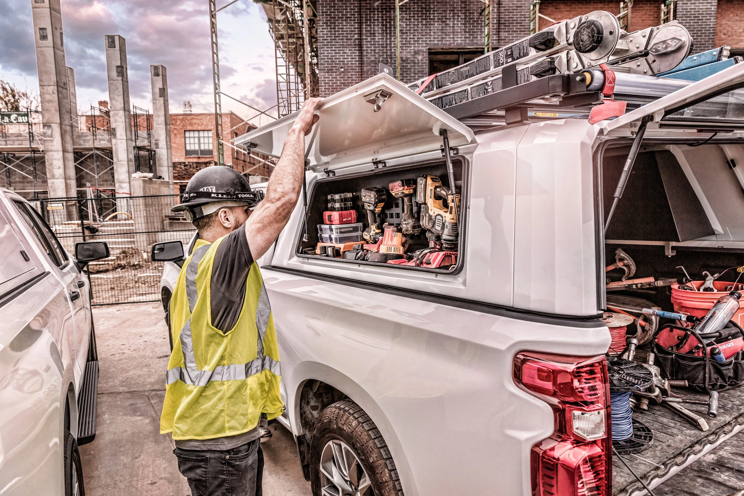 Construction worker in a yellow safety vest and helmet opening a toolbox in the back of a white pickup truck at a construction site.