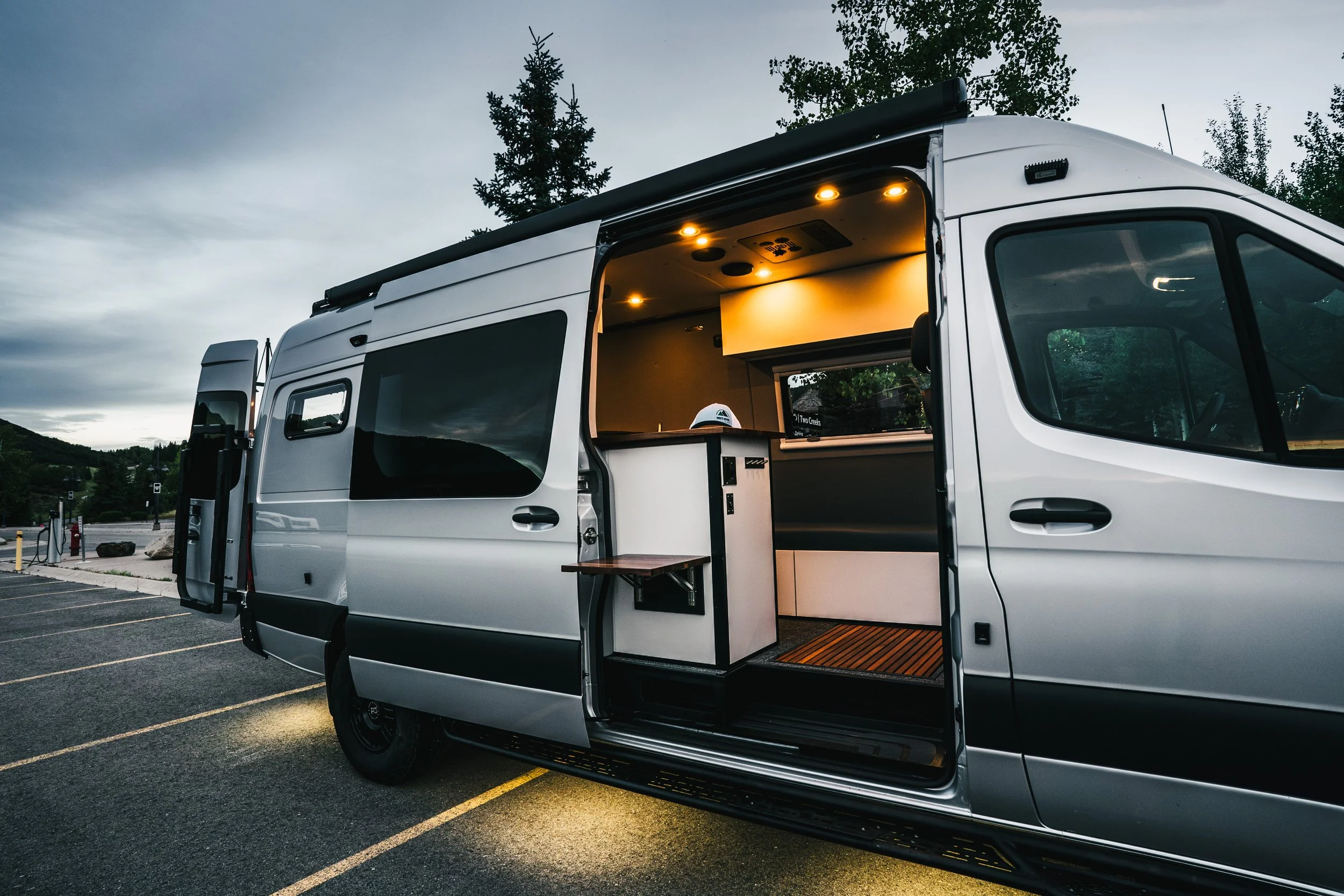 A silver camper van parked in a parking lot with a view of trees and hills in the background. The side door is open, revealing a lit interior with storage cabinets and a small counter.