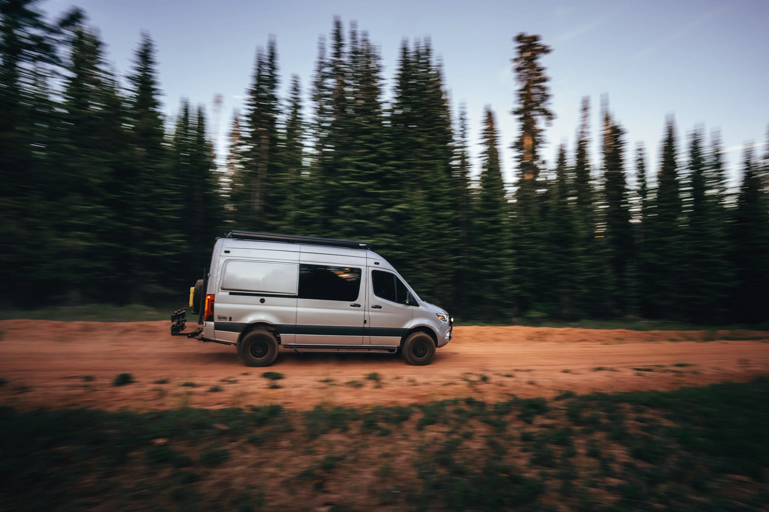 A silver camper van driving on a dirt road in a forest.