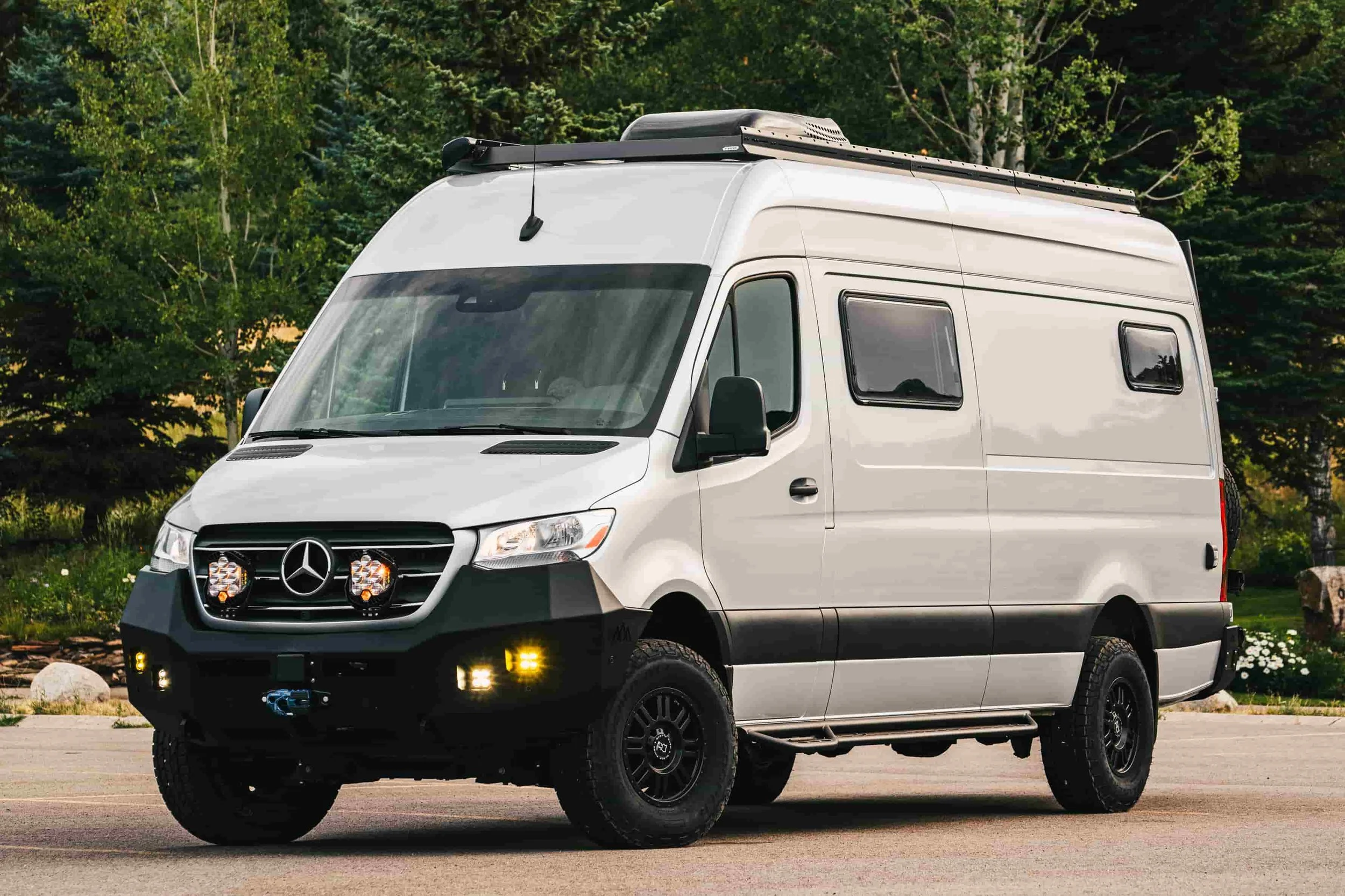 Silver Mercedes-Benz camper van parked on a paved road with a green, wooded background.