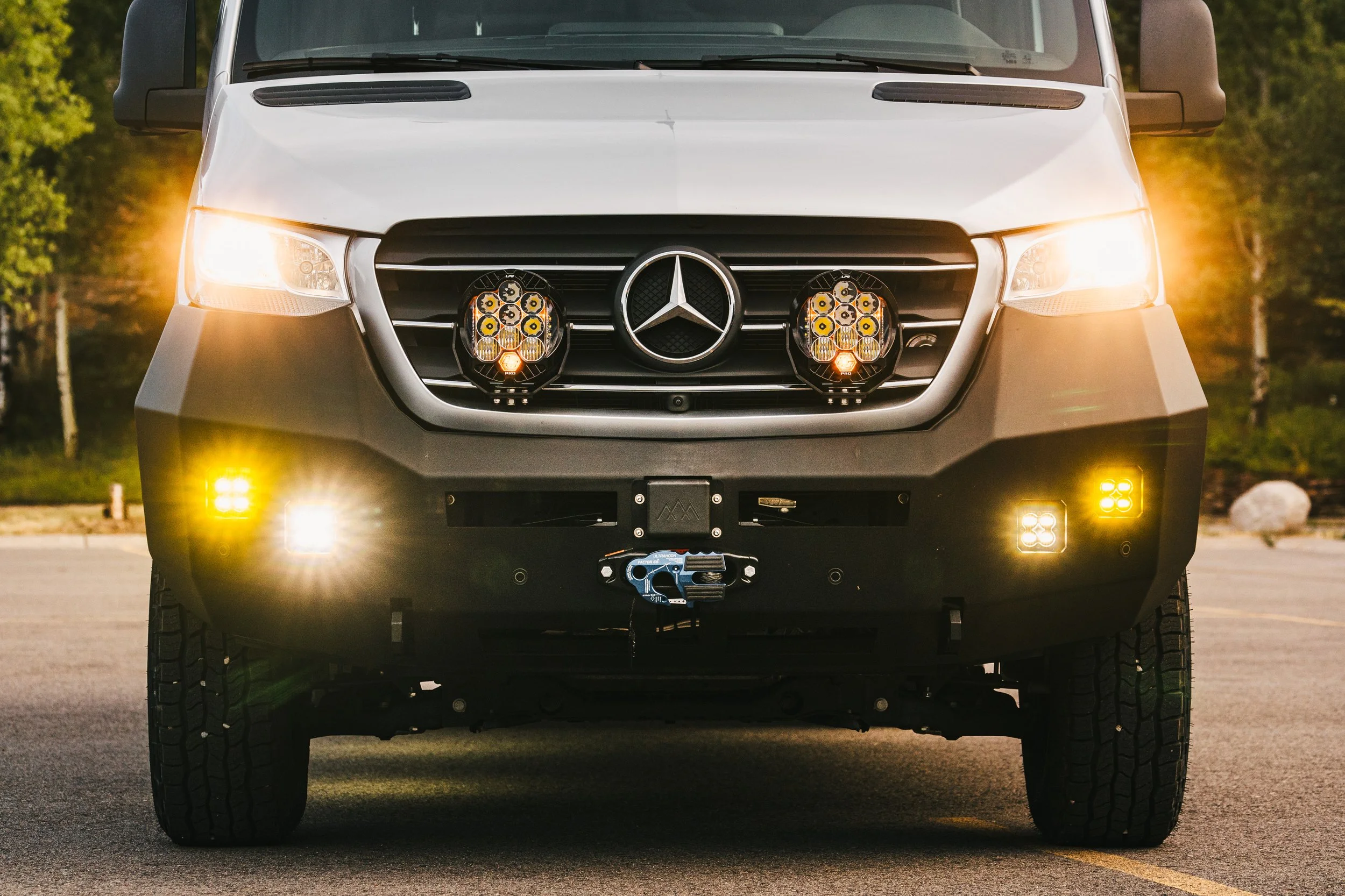 Front view of a Mercedes-Benz Sprinter van equipped with off-road accessories, including additional lights on the grille, fog lights on the bumper, and a winch hook, parked on a paved surface with trees in the background.