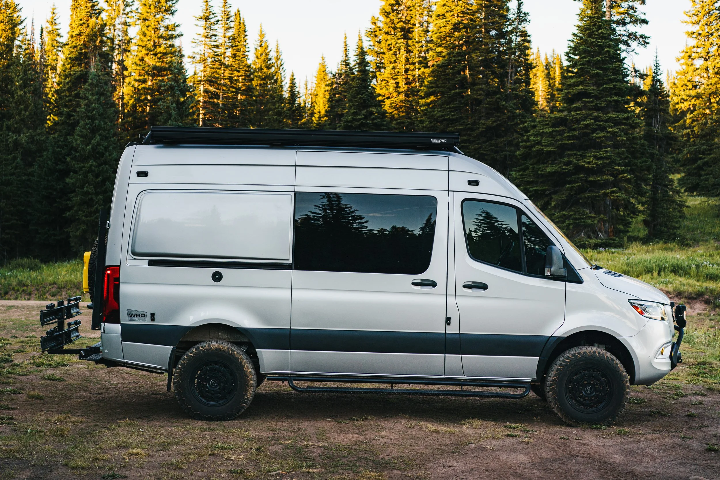 A white camper van parked on a dirt area surrounded by a forest of tall pine trees.