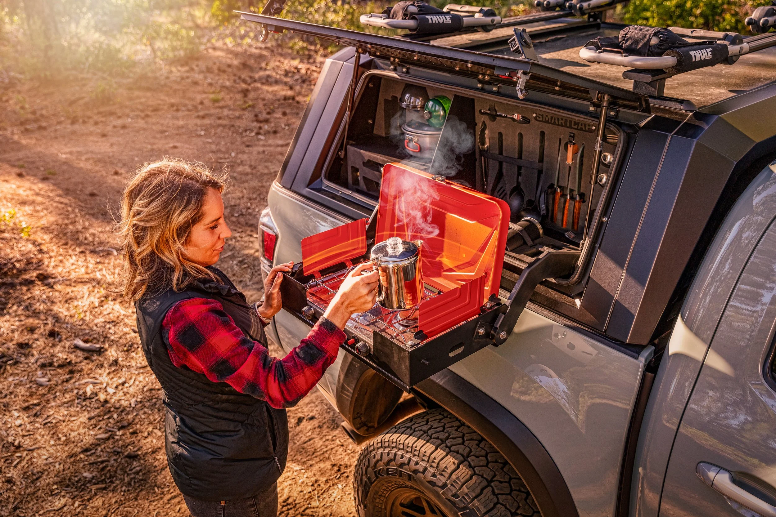 A woman in outdoor gear prepares tea with a kettle on a portable stove from a mounted rear outdoor kitchen on a vehicle, in a wooded outdoor setting during sunlight.