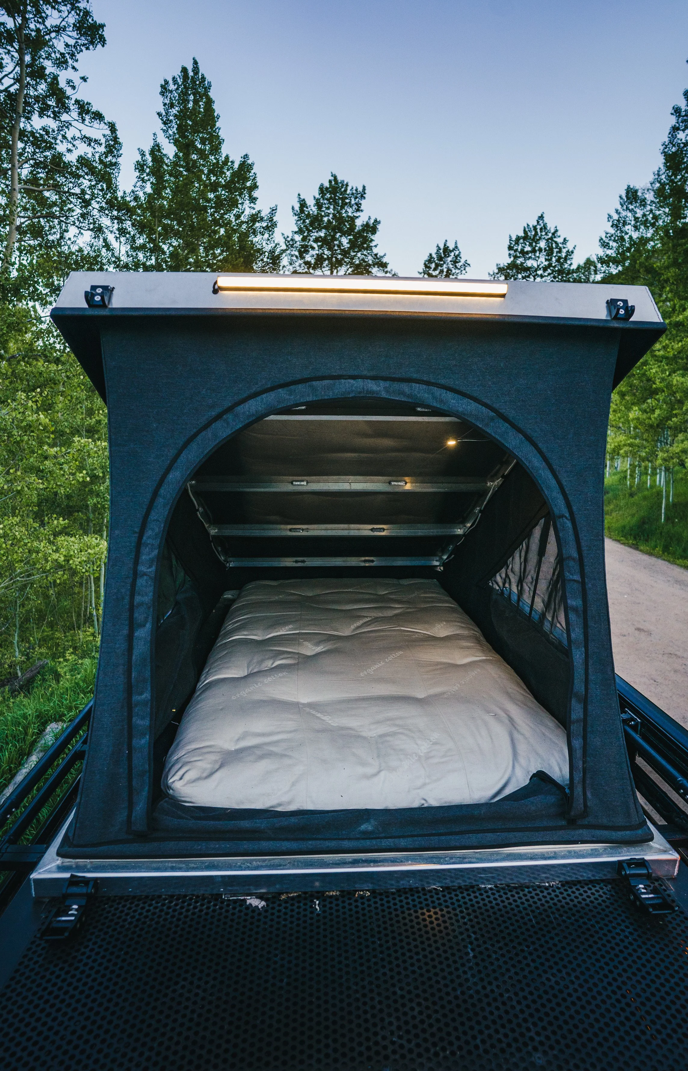 A small camping tent with an open roof, revealing a cushioned mattress inside, set up outdoors on a dirt path surrounded by green trees during twilight.