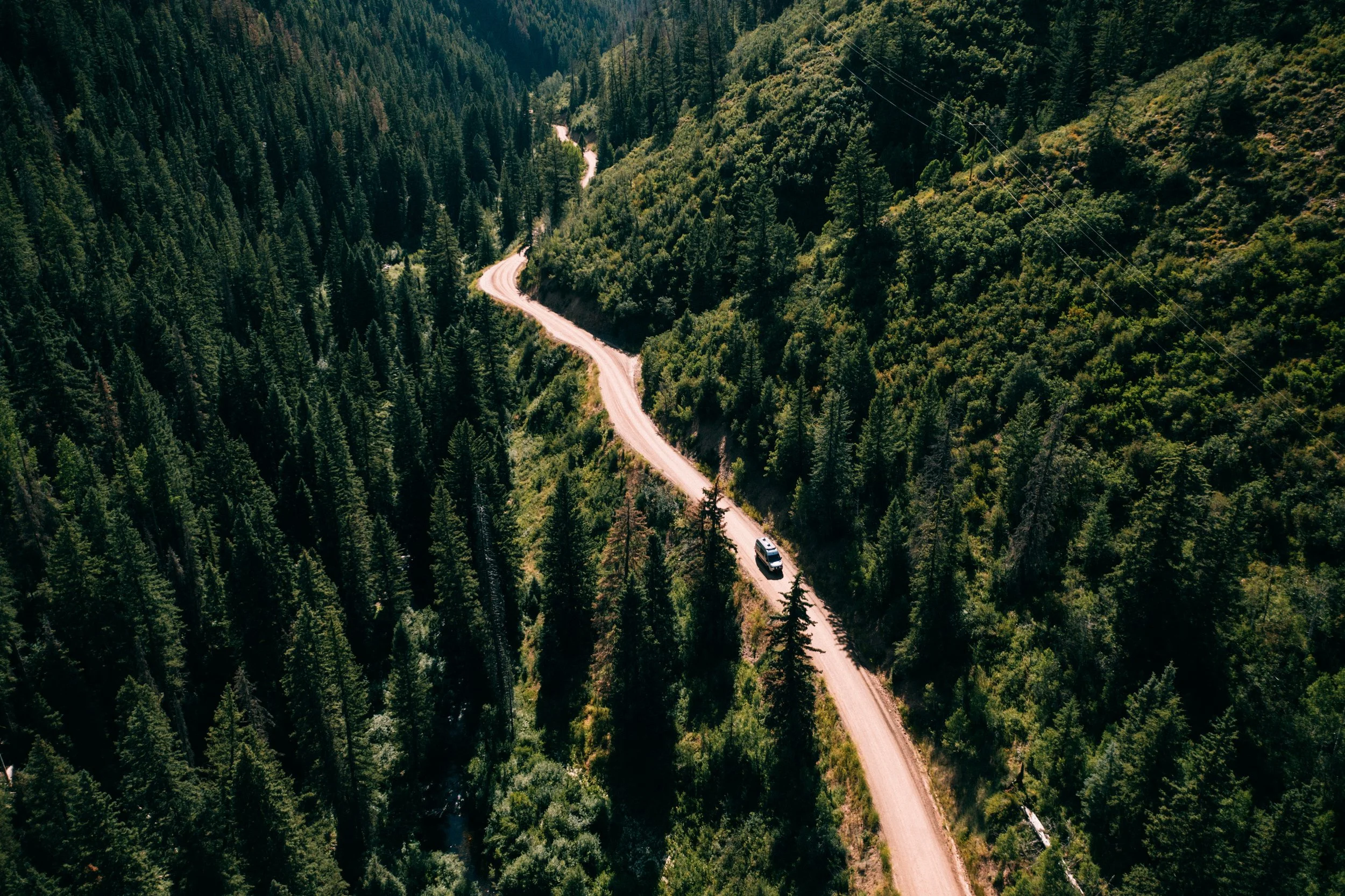 An aerial view of a winding dirt road through a dense forested mountain area with a single vehicle traveling along the road.