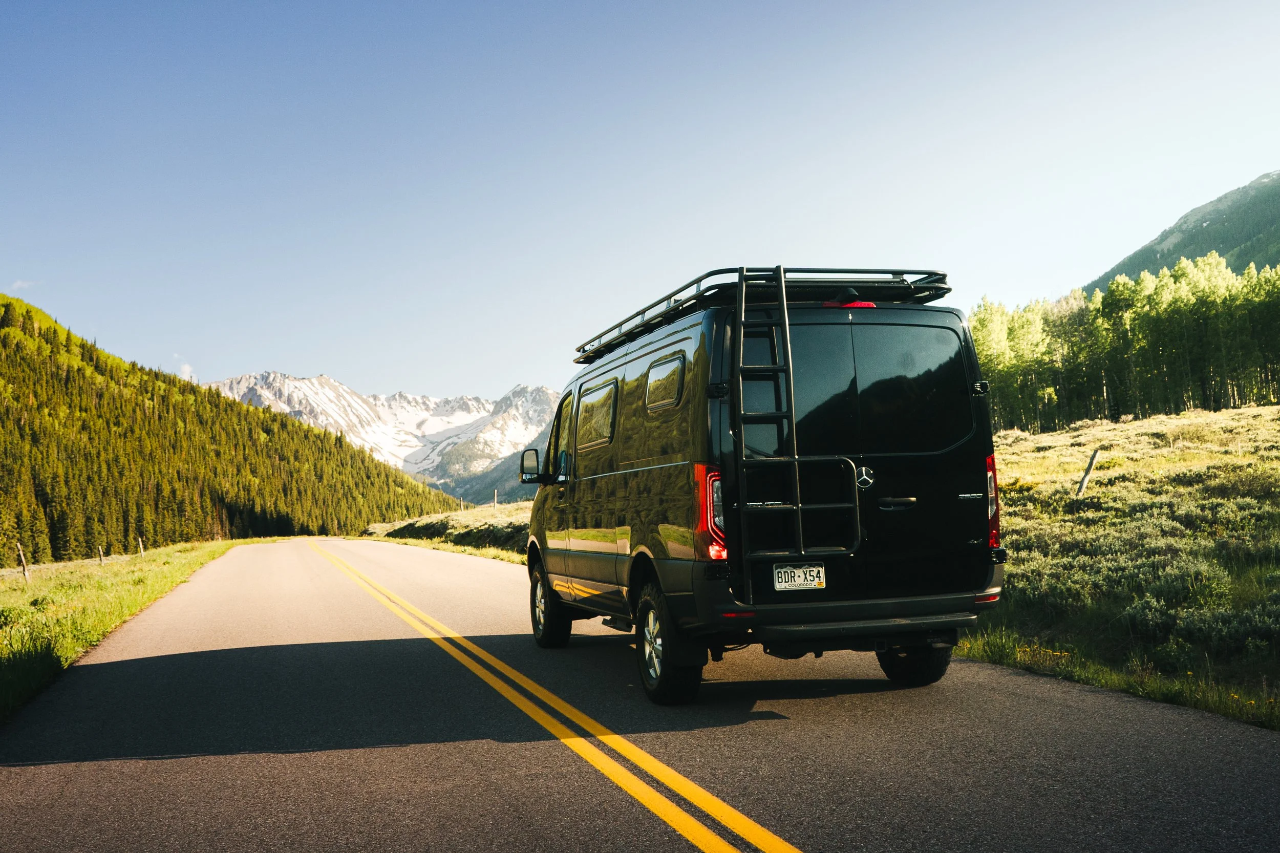 Black van parked on a two-lane road in a mountainous area with green trees and snow-capped peaks in the background.
