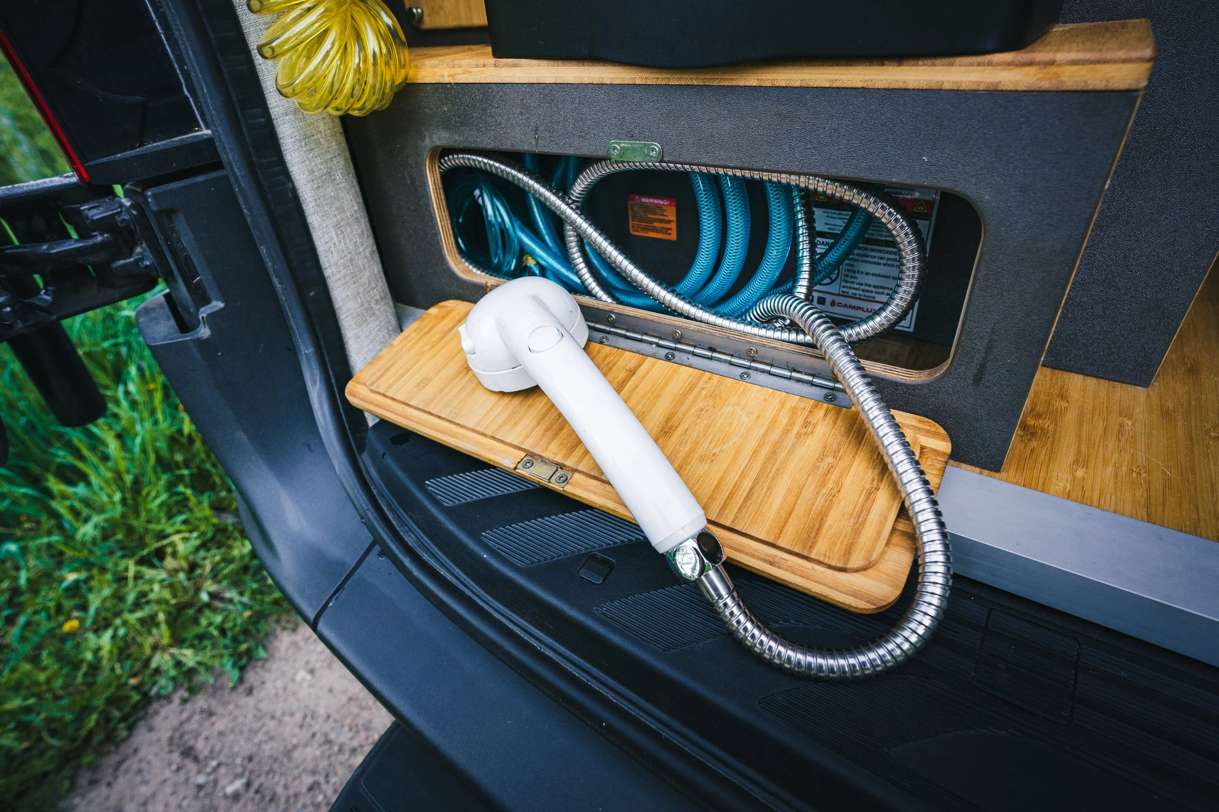 A portable vacuum cleaner with a white nozzle and a flexible hose resting on a wooden surface, next to a compartment filled with blue and silver cables inside a camper van or small RV.