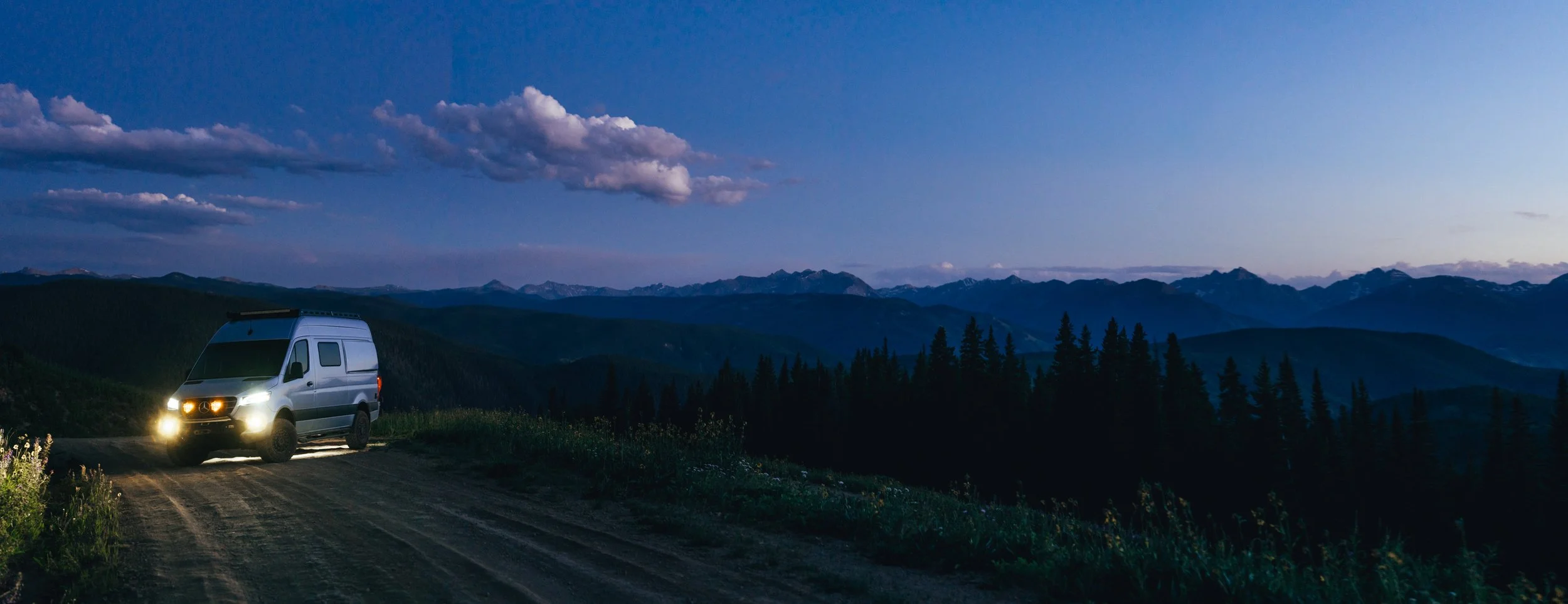 A camper van parked on a dirt road in a mountainous landscape during twilight with a sky filled with clouds and distant mountain peaks.