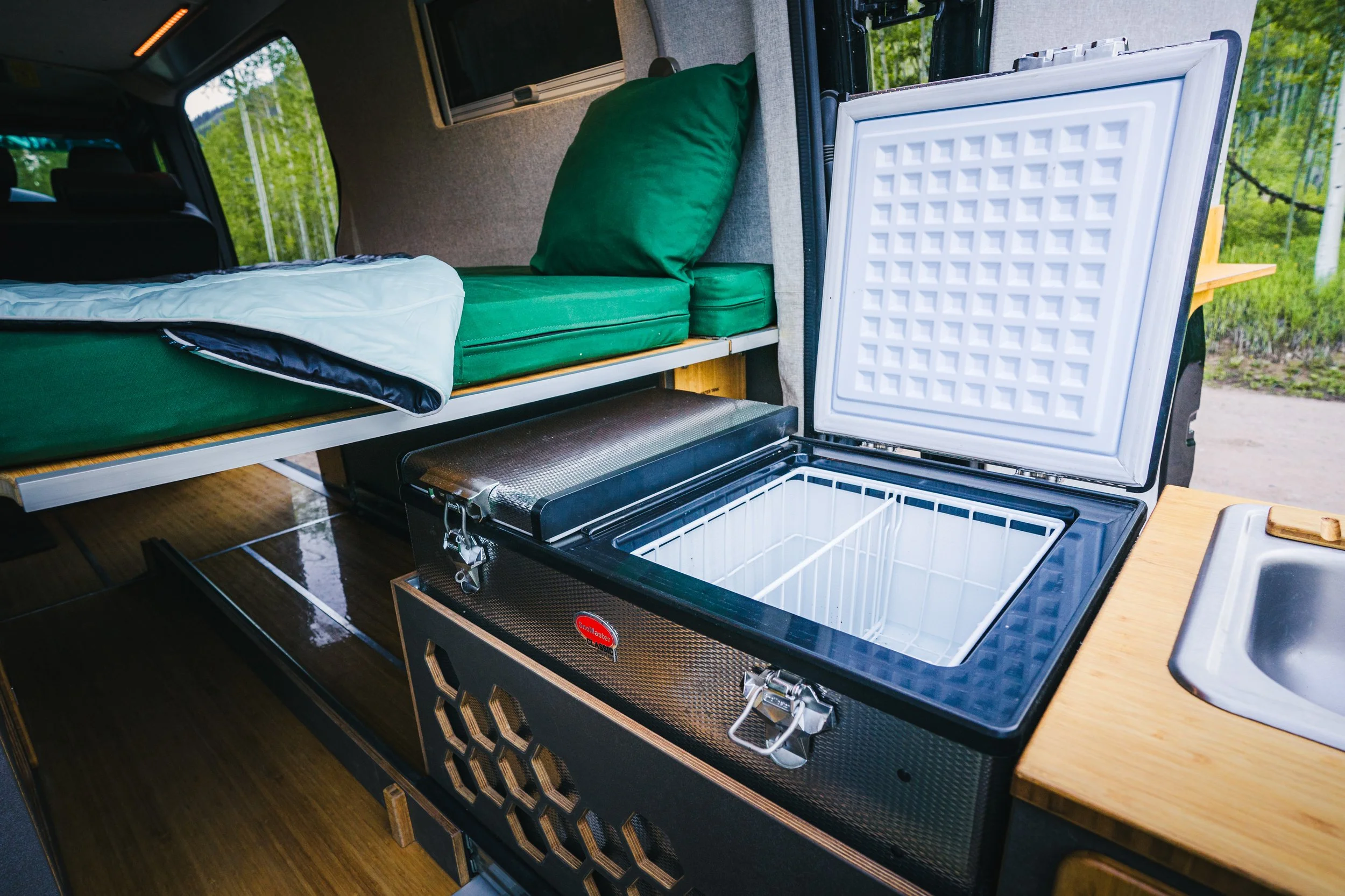 Inside a camper van showing the open fridge, bed with green bedding, and a window with greenery outside.