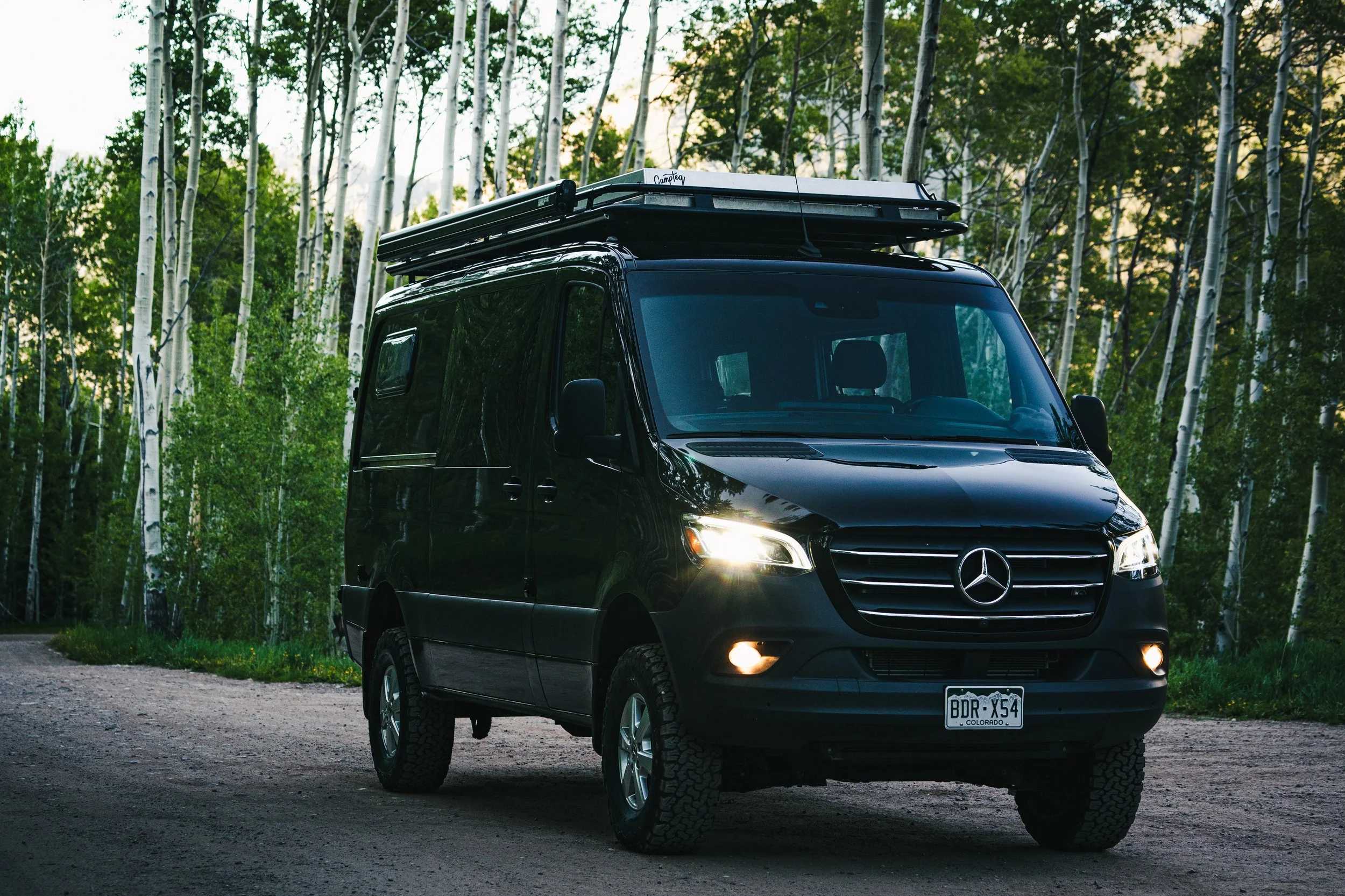Black Mercedes-Benz camper van parked on a gravel road in a forest, with trees and green foliage in the background, and the van's headlights and fog lights turned on.