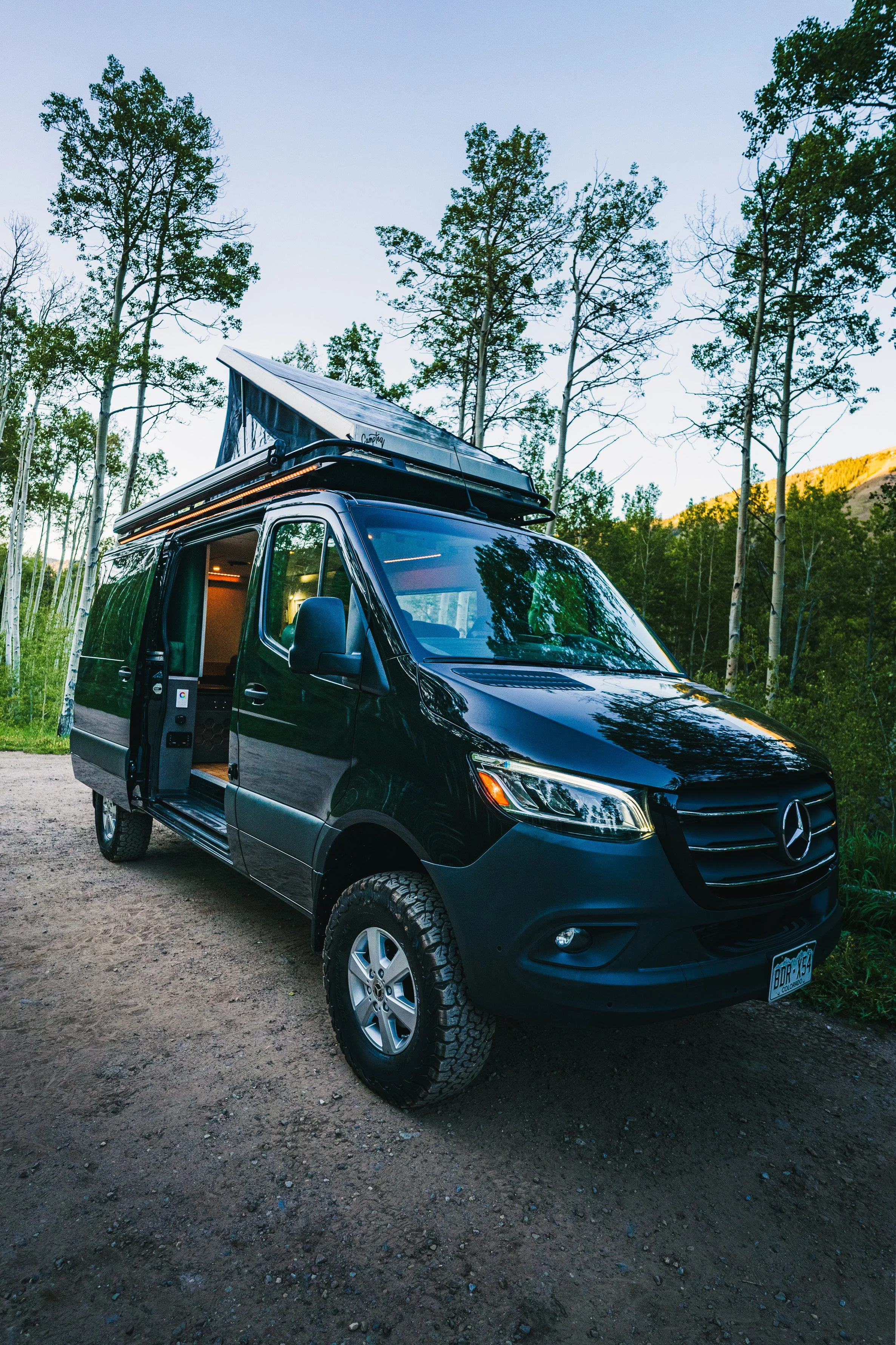Black camper van with a pop-up roof, parked on a dirt trail surrounded by tall trees in a forested area during daytime.