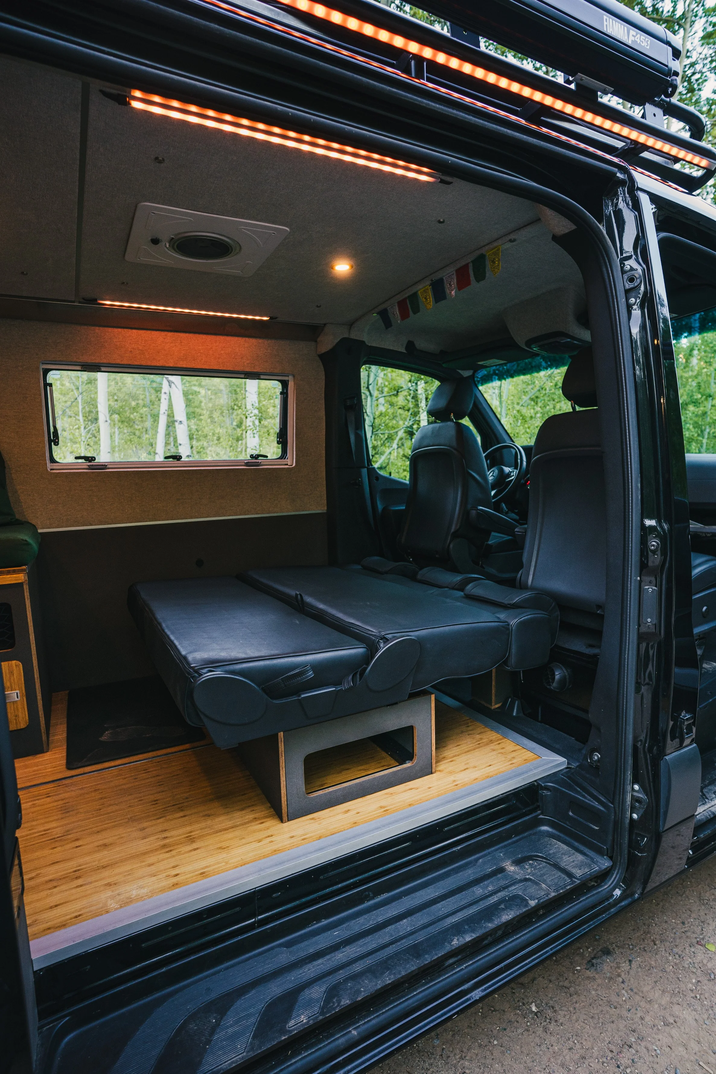 Interior of a camper van with a foldable bed, black leather seats, wood flooring, and a window showing trees outside.
