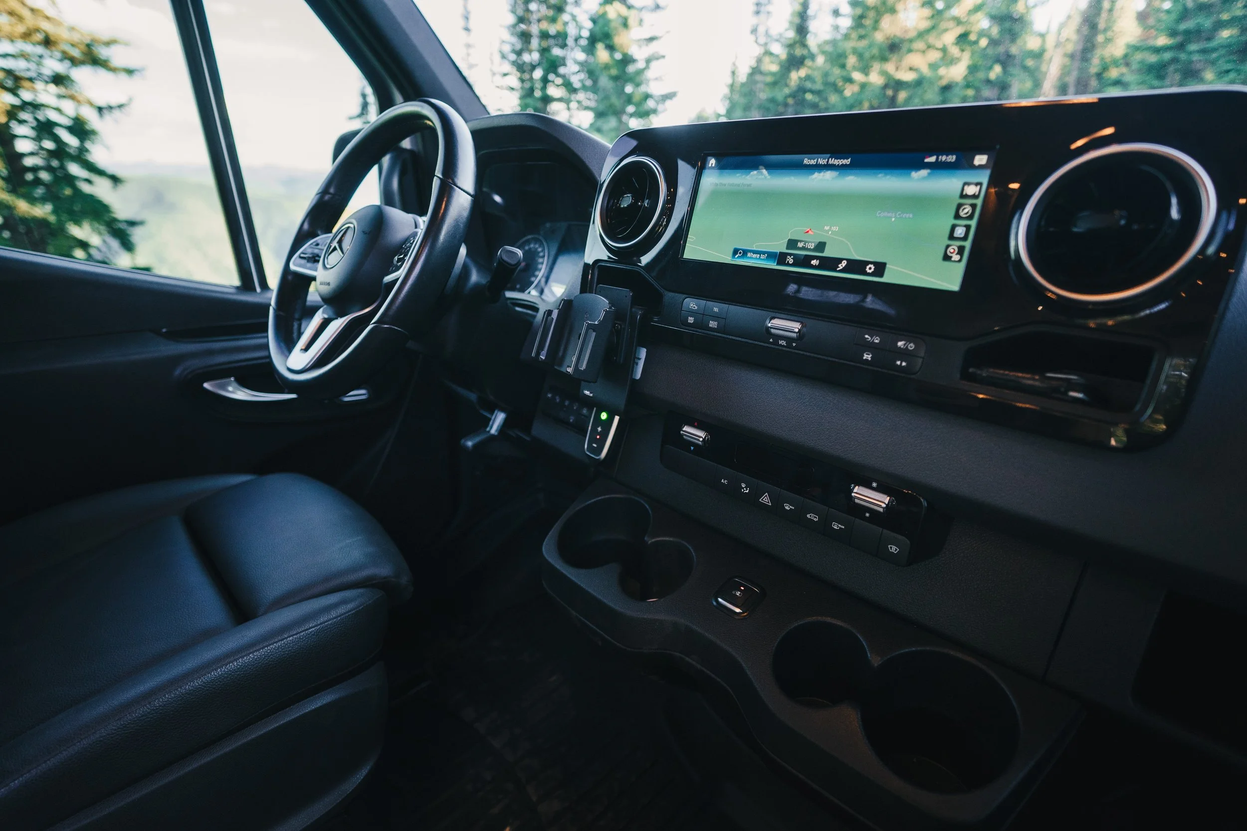 Interior of a Mercedes-Benz van showing the dashboard, steering wheel, and infotainment screen with navigation, surrounded by trees visible through the windshield.