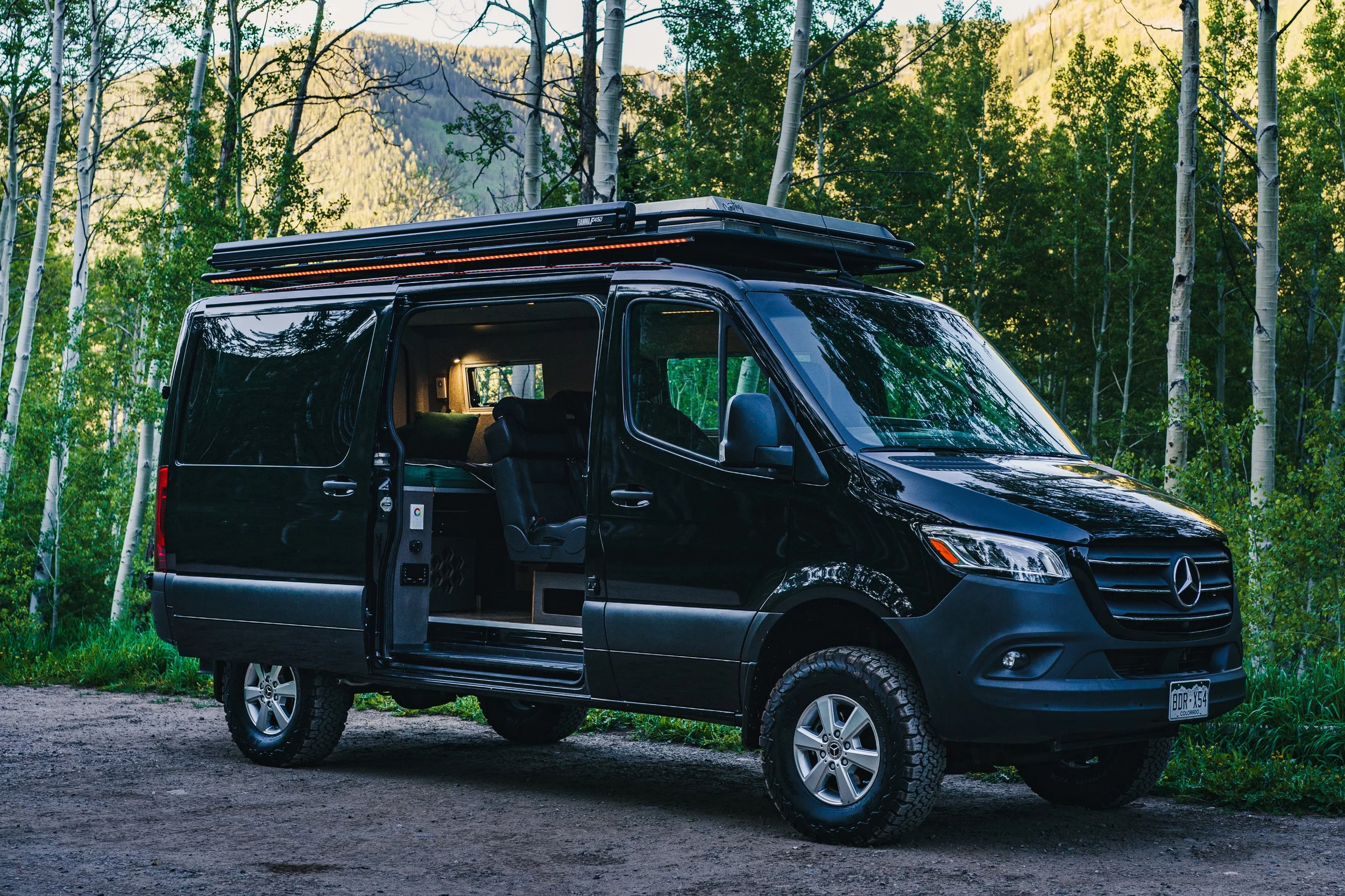 Black camper van parked in a wooded area with trees and mountains in the background, with an open sliding door revealing the interior.