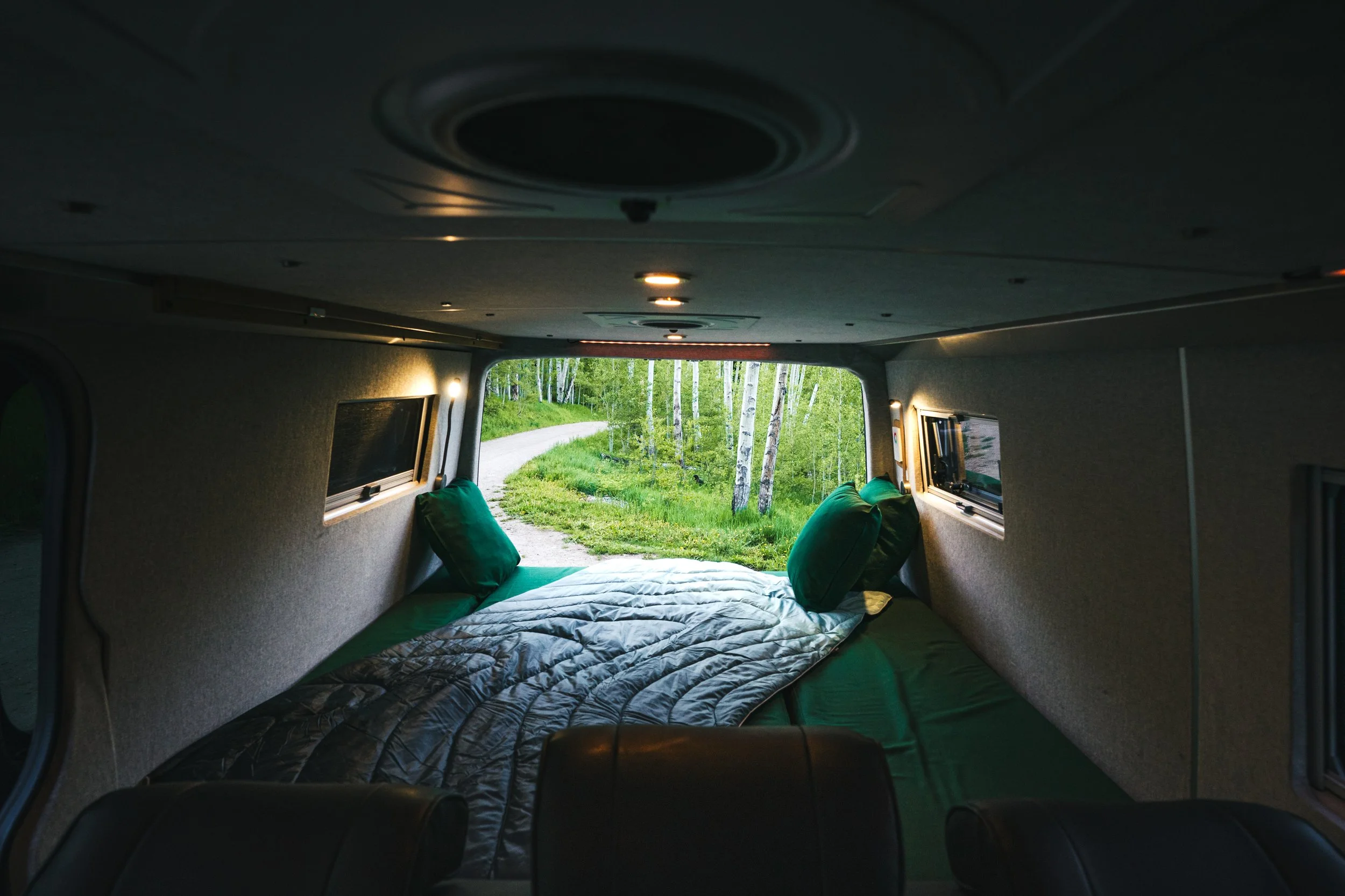 Interior of a campervan with bed facing windows showing a forest and dirt road outside.