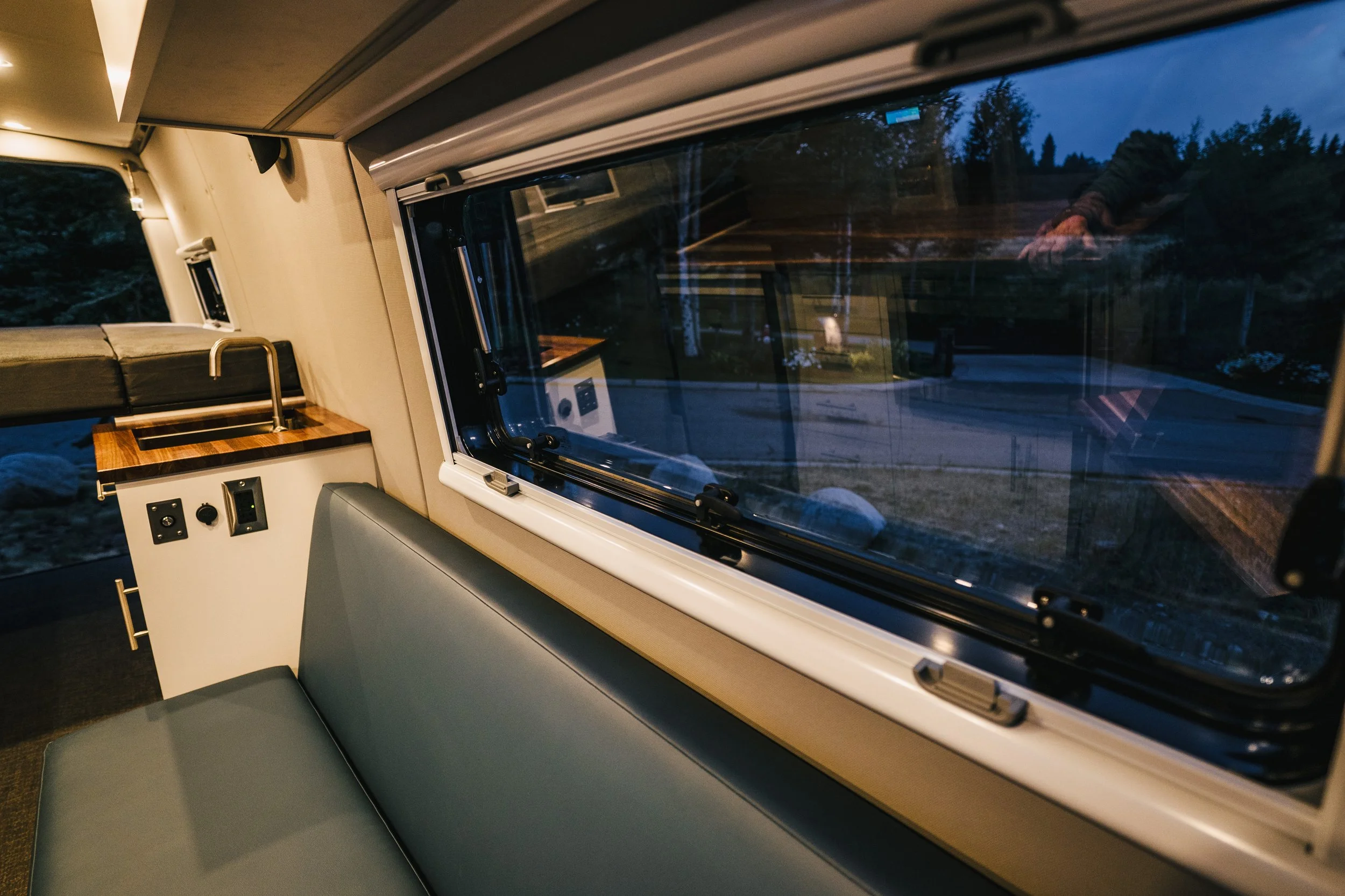 Interior of a camper van with a large window showing an outdoor landscape at dusk. The interior features a bench seat, electrical outlets, and a small wooden countertop with a sink.
