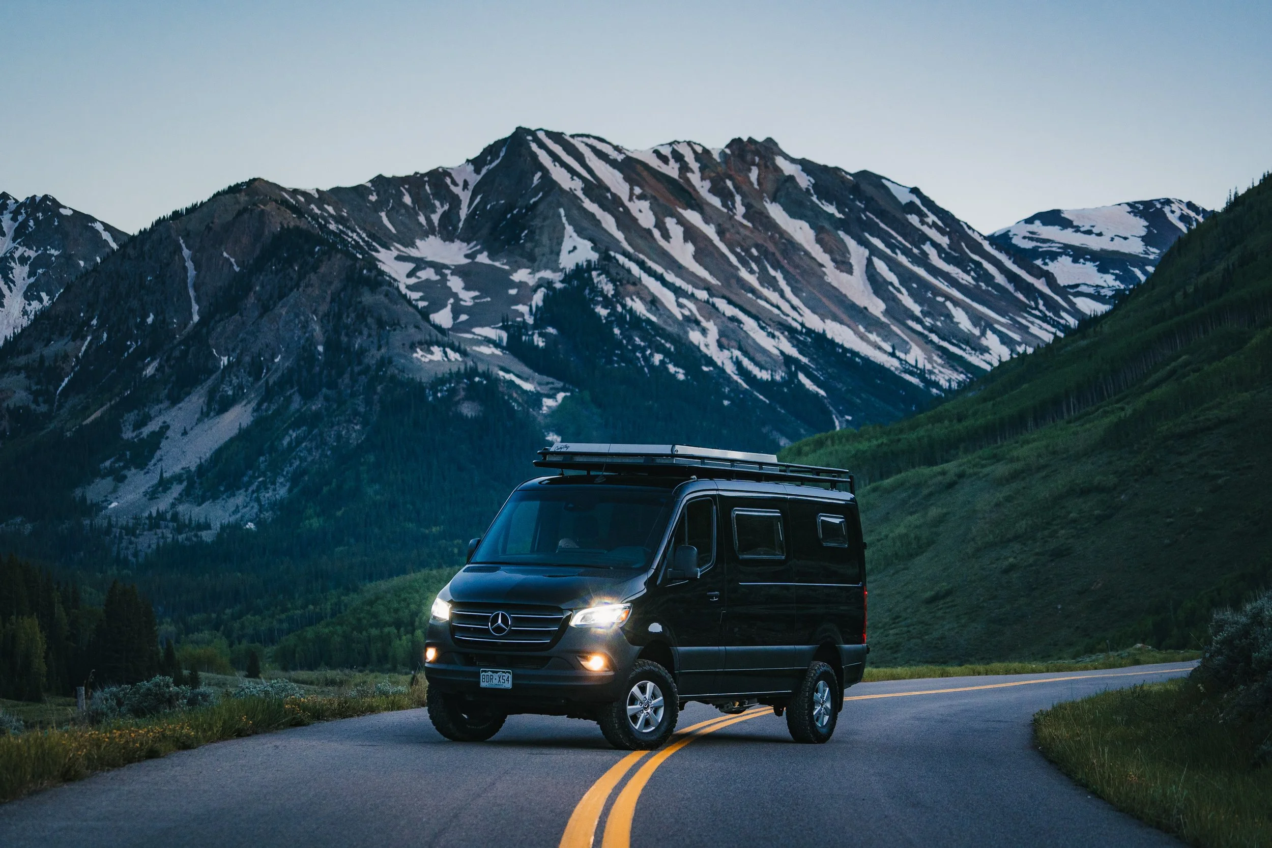 Black van parked on a winding mountain road, with snow-capped mountains and green forested hills in the background.