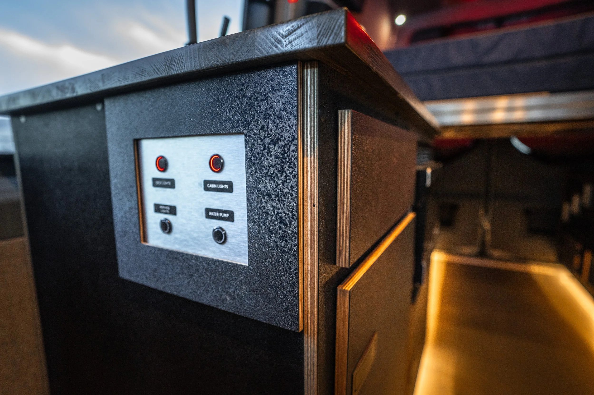 Close-up of a black control panel with switches labeled 'Indoor/Outdoor,' 'Cabin Lights,' and 'Water Pump' on a black textured surface next to a dark wood table or countertop.