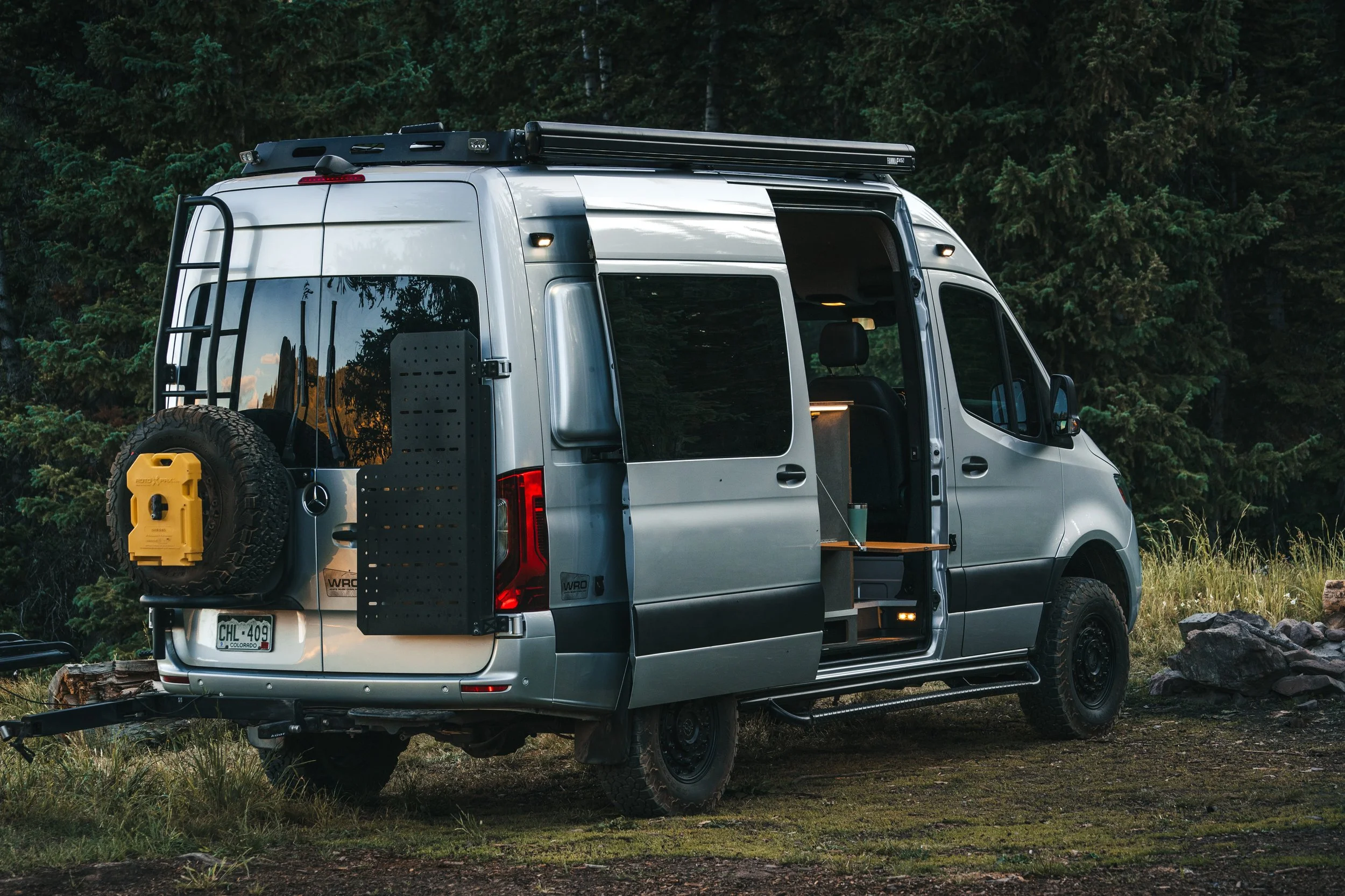 Silver camper van parked outdoors in a forested area, with a spare tire mounted on the back and a yellow container attached to it, and an open side door showing a small interior space.