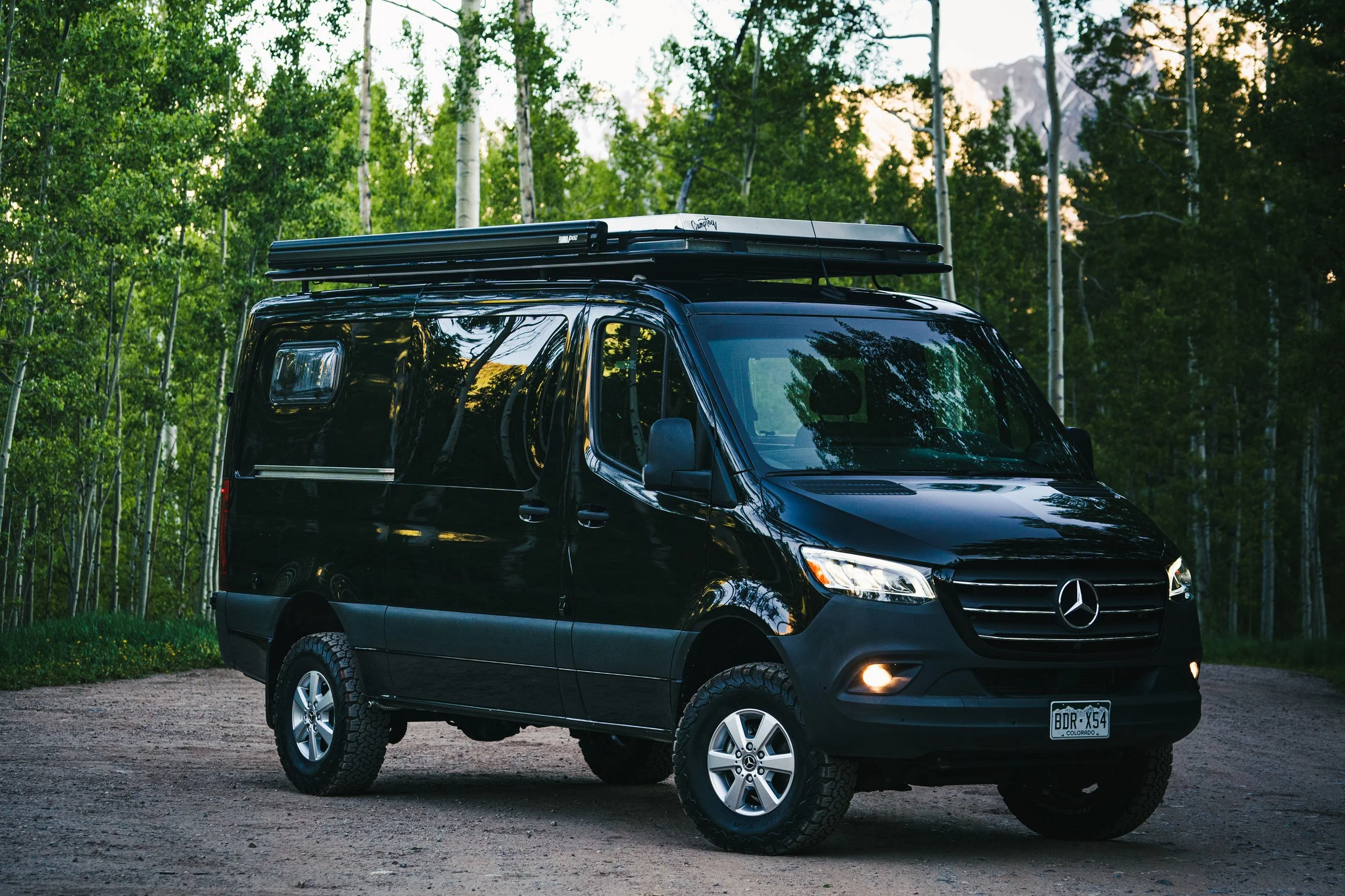 A black Mercedes-Benz camper van parked on a dirt road in a forest with tall trees and mountains in the background.