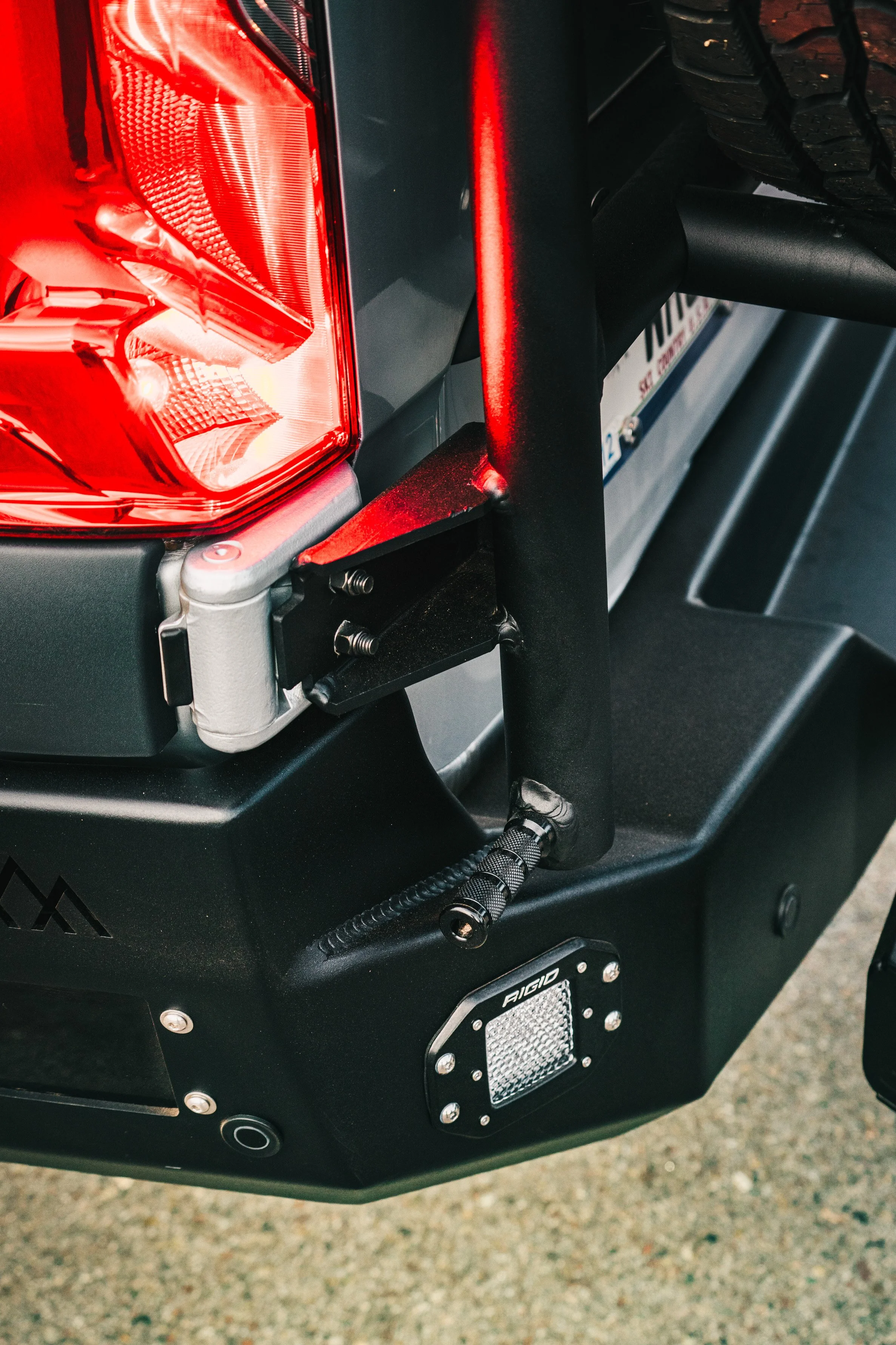 Close-up of the rear corner of a vehicle, showing a red tail light, black bumper, and part of a tire, with a black suspension component and a small LED light array labeled 'Aigid'.