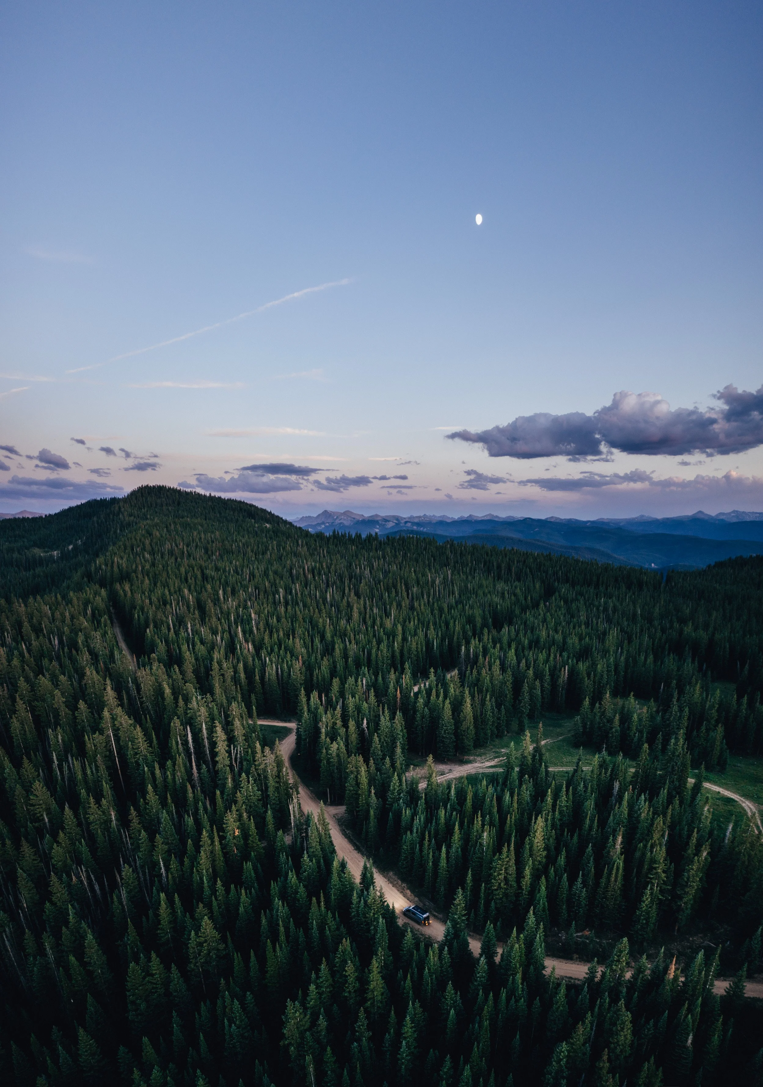 Aerial view of a forested mountain landscape under a cloudy sky with a visible moon and a winding dirt road with a vehicle.
