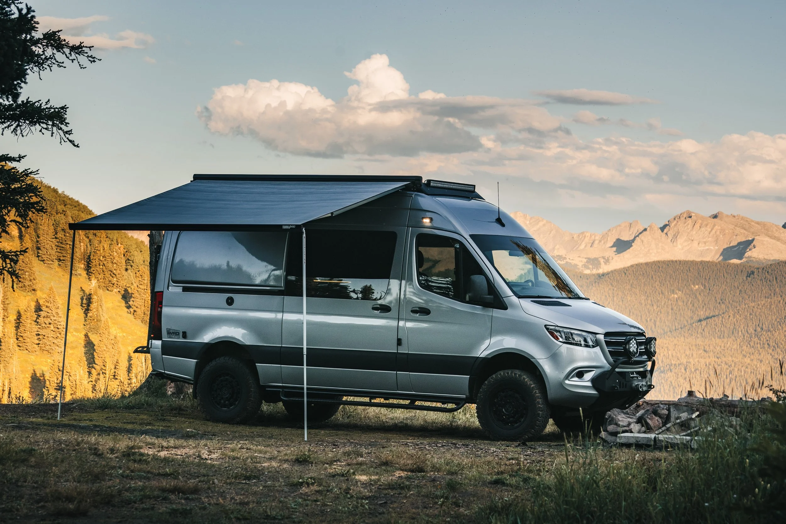 Silver camper van outdoors with an awning, mountain range in the background, trees and cloudy sky.