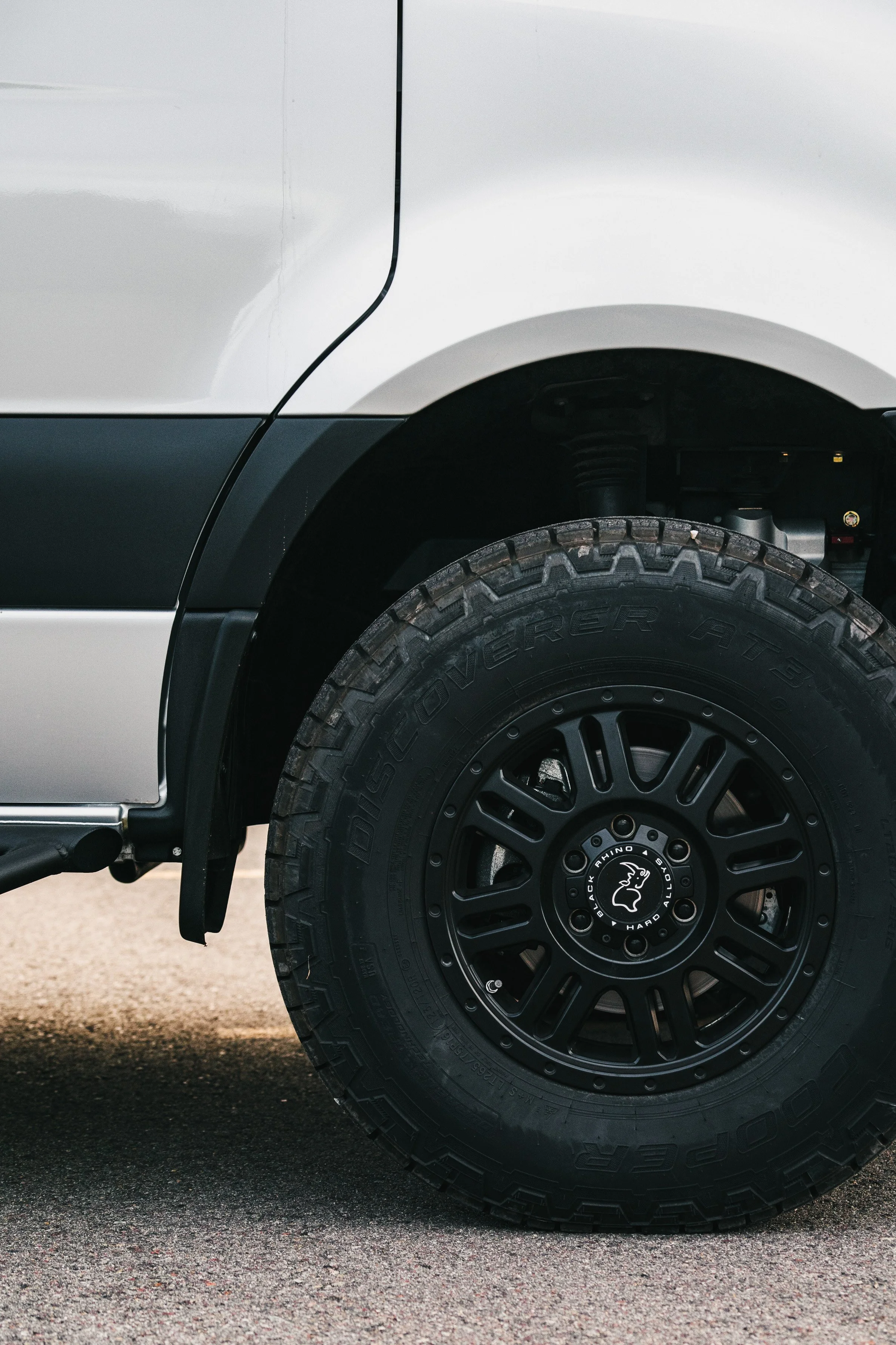 Close-up of a black off-road tire mounted on a black rim, attached to a white vehicle, with a visible suspension component.