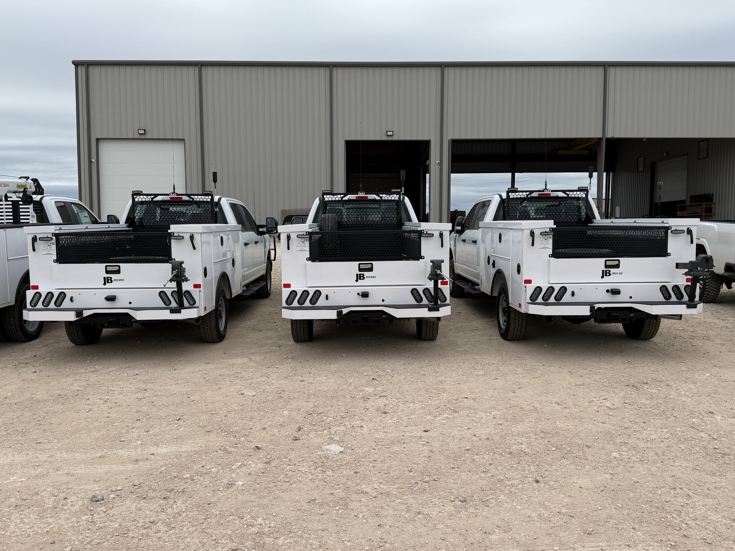 Three white utility trucks with flatbeds and black protective guards parked in front of a large gray industrial building.