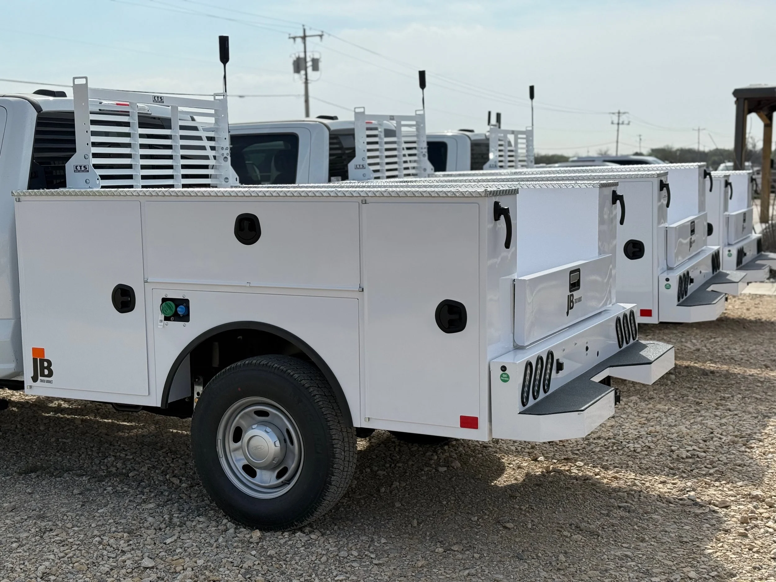 A row of white utility trucks with compartments and steps on a gravel lot, with utility poles and wires in the background.