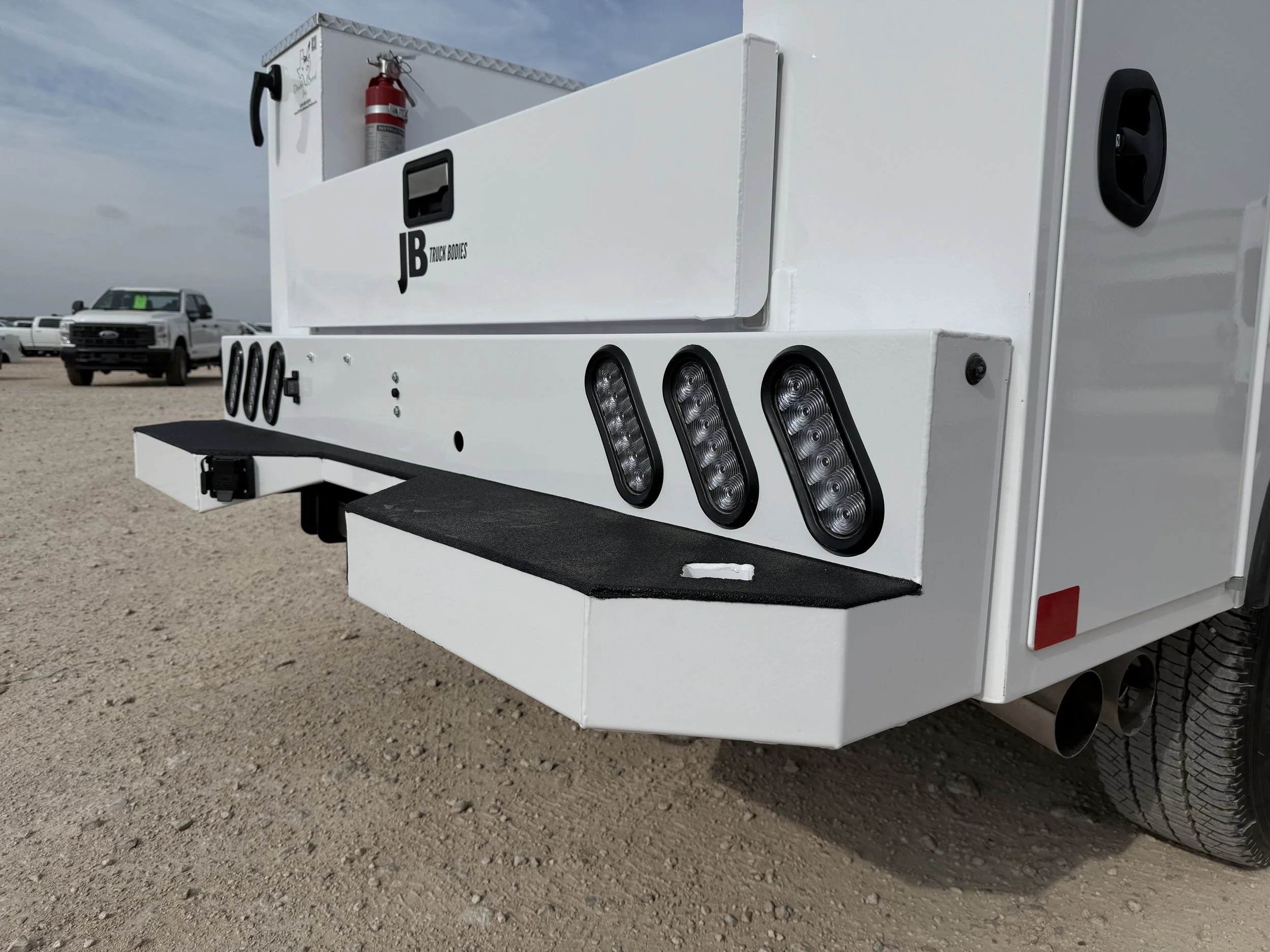 Close-up of the back of a white utility or service vehicle with LED lights and a fire extinguisher mounted on top, parked on a dirt lot with other trucks in the background.