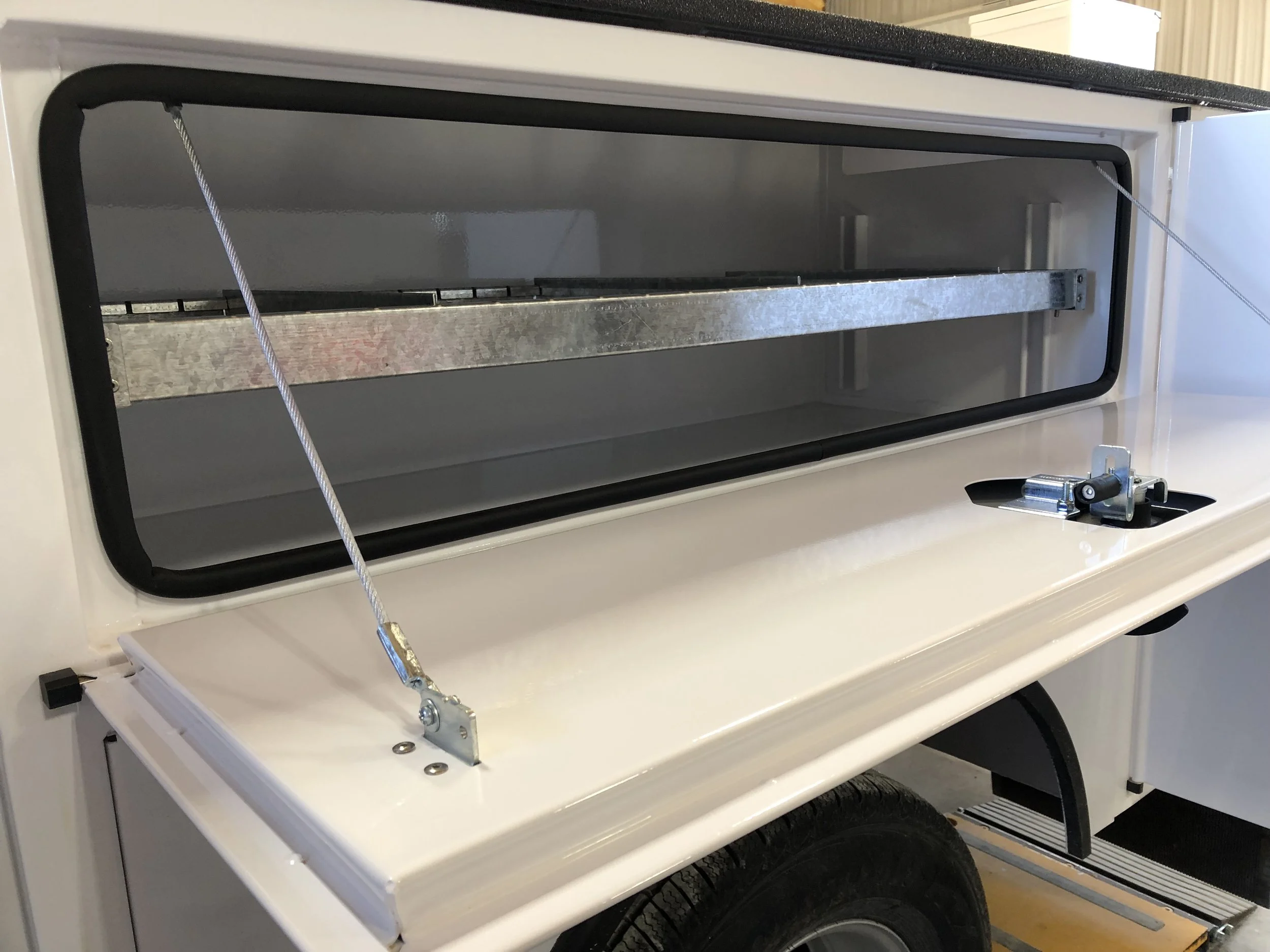 Close-up of a white trailer with an open hatch and a metal shelf inside, secured with a cable and locking mechanism.