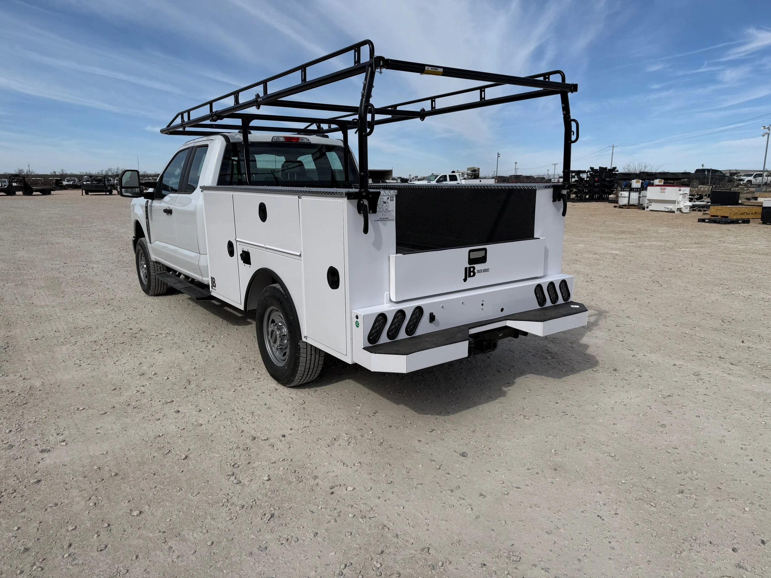 White utility truck with black equipment rack on top and storage compartments on the side, parked on a dirt lot under a blue sky.