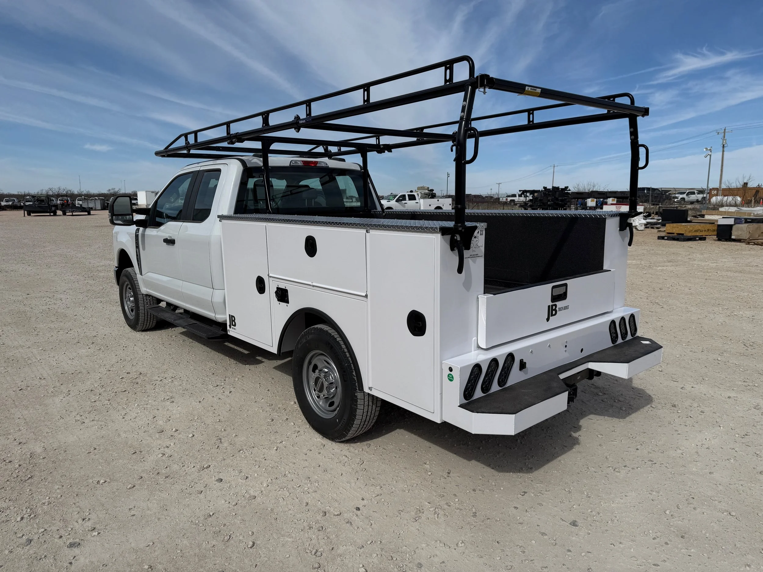 White service pickup truck with black utility racks and storage compartments parked on a gravel lot under a partly cloudy sky.