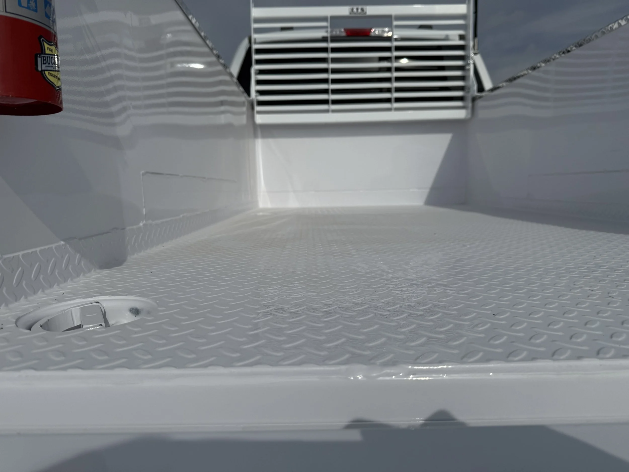 The image shows the interior of a flatbed truck or trailer, with a white textured metal surface, a drain hole in the corner, and a side wall with a fire extinguisher mounted on the left side. The front has a grille or vent for airflow.